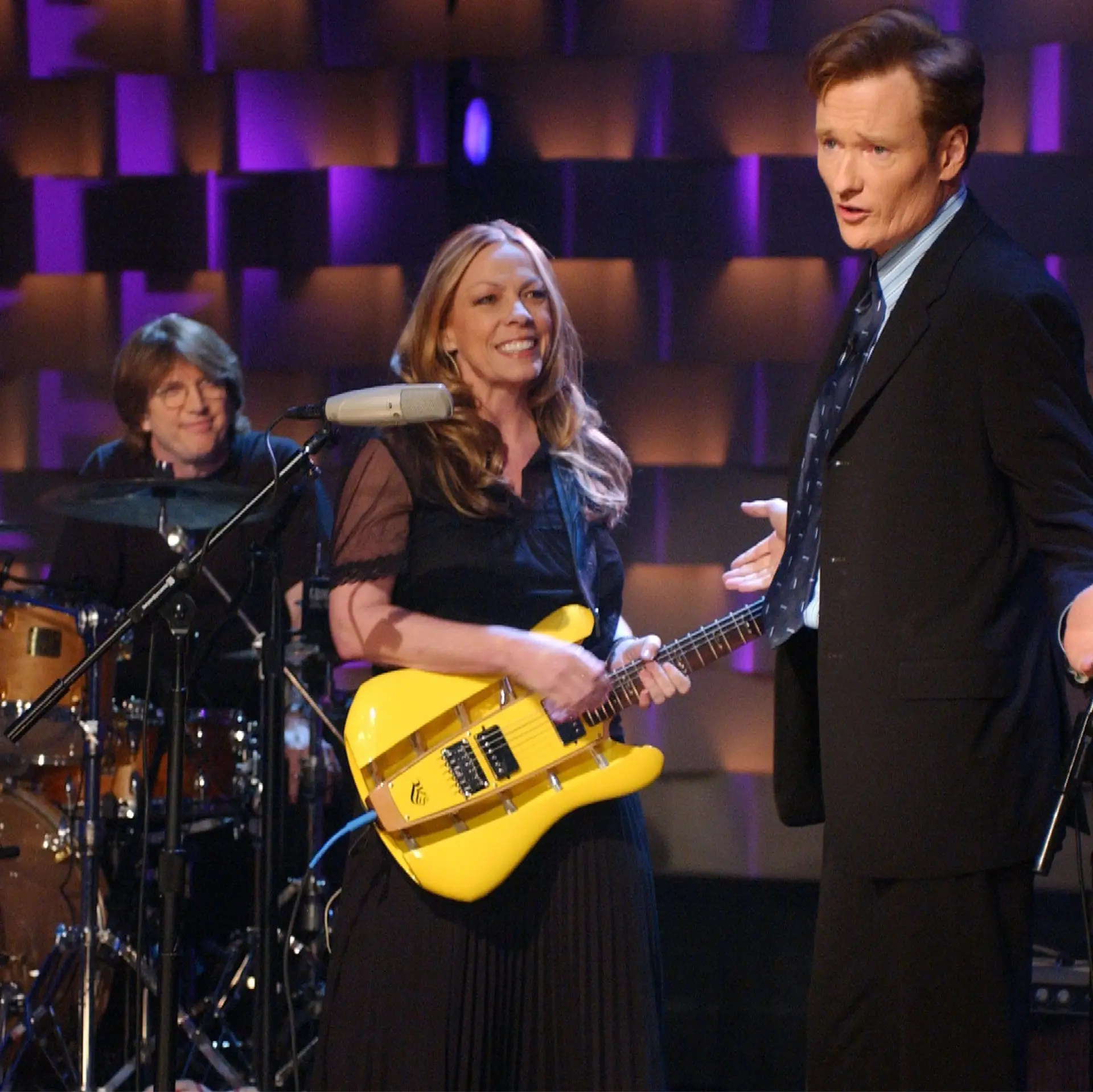 Smiling woman plays a yellow RKS guitar on a TV stage beside a man in a suit, with a drummer behind her