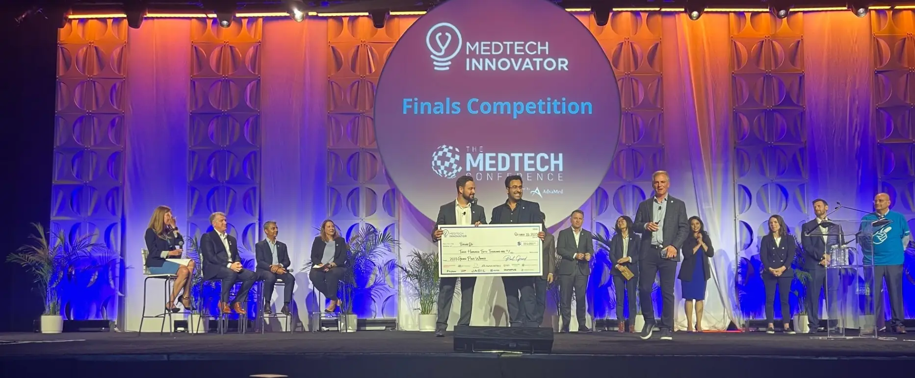 A group of people on stage at the MedTech Innovator Finals Competition, with a large…