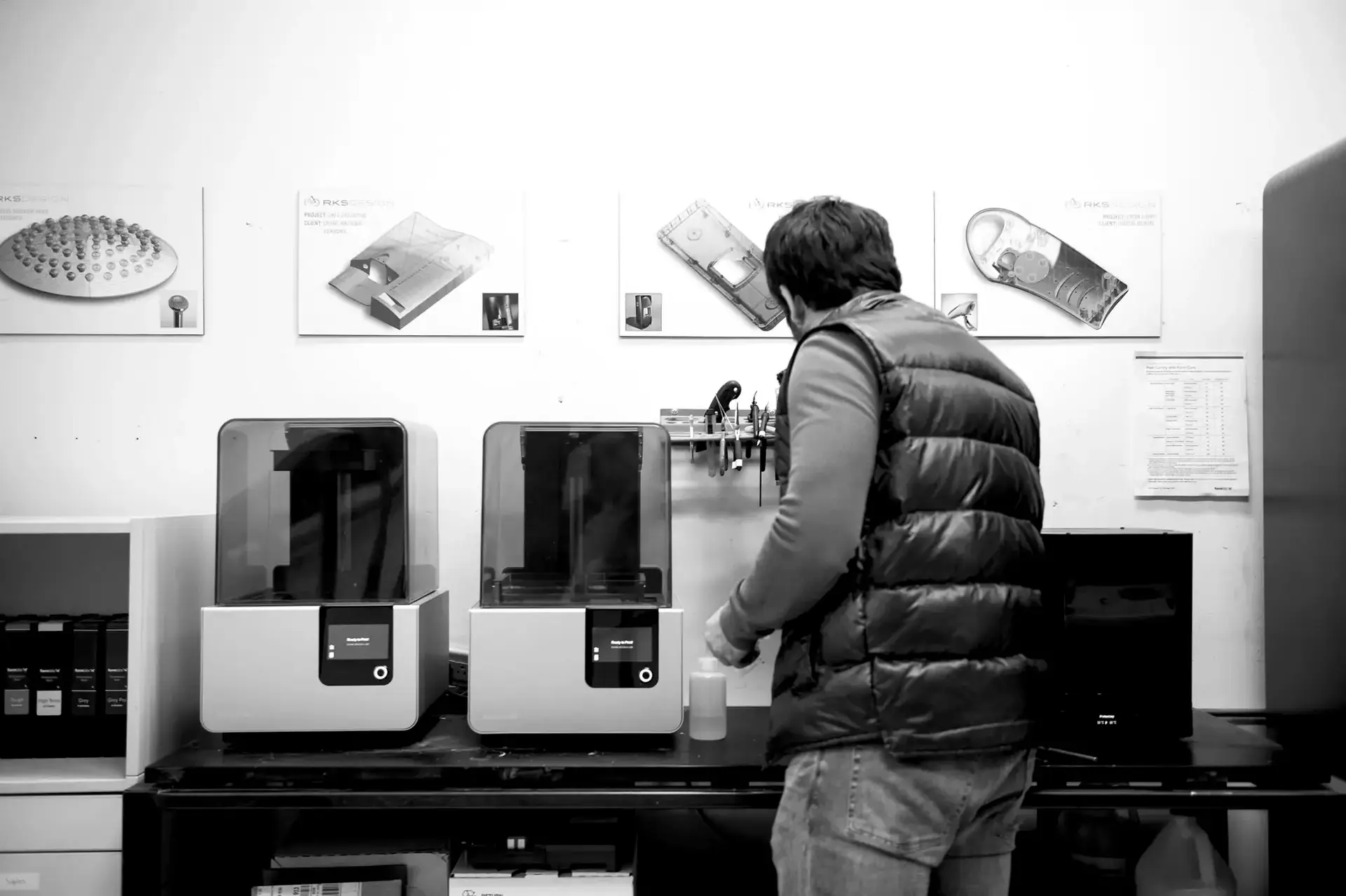 A person wearing a puffer vest operates 3D printers on a workbench in a lab.