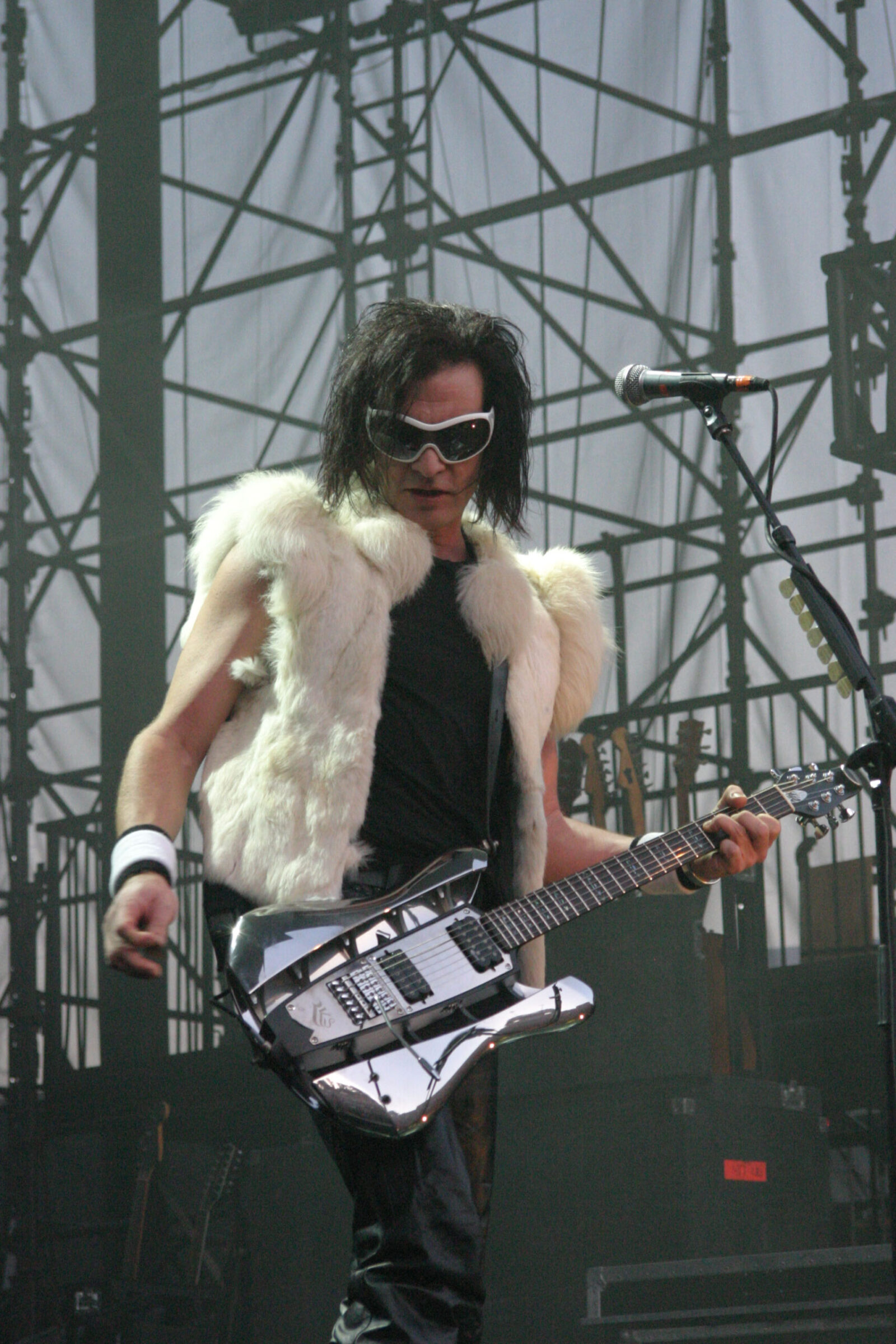 A guitarist in sunglasses and a white fur vest plays a chrome-finished RKS guitar on an outdoor stage