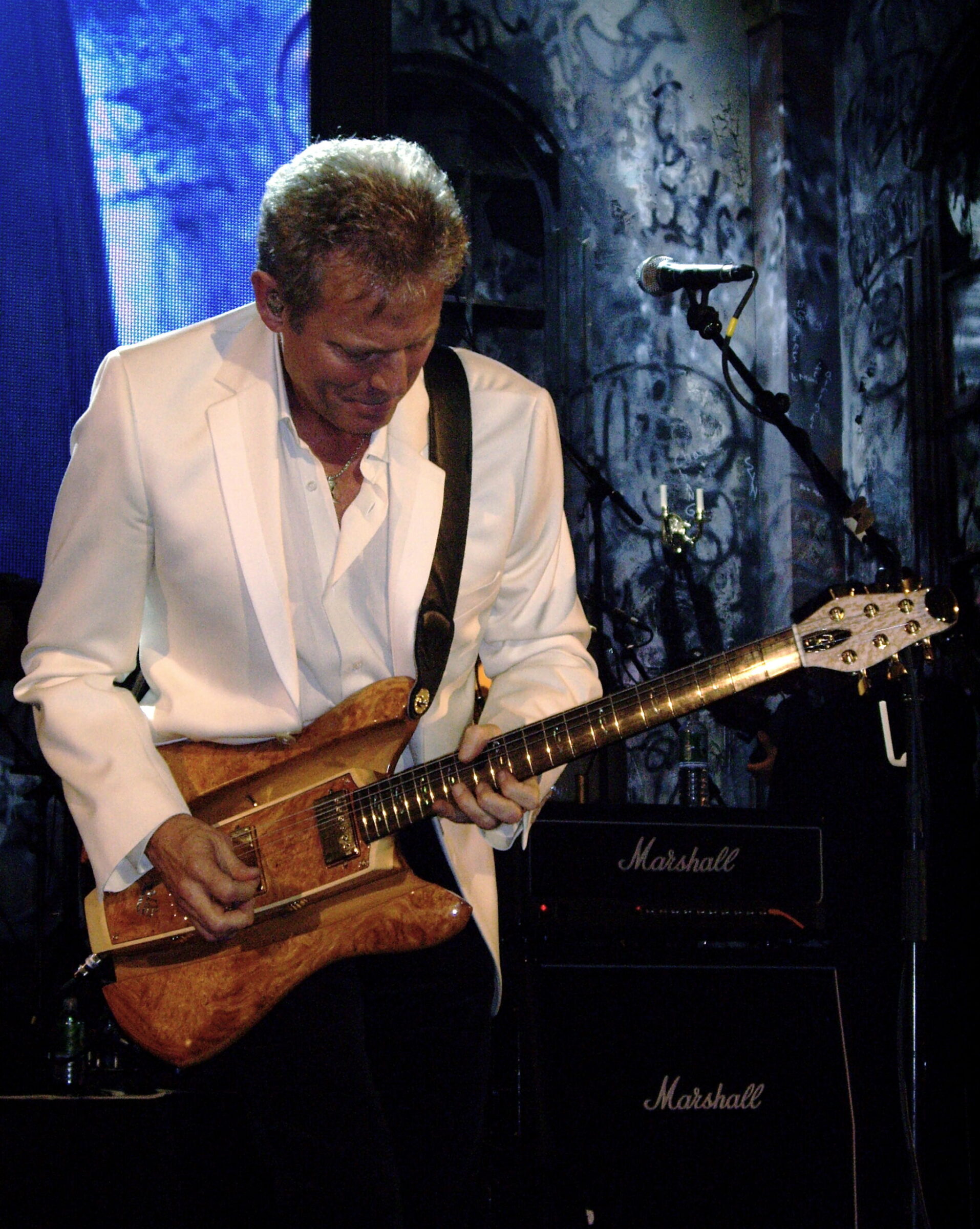 A guitarist in a white blazer plays a burl-wood RKS guitar on stage in front of Marshall amplifiers