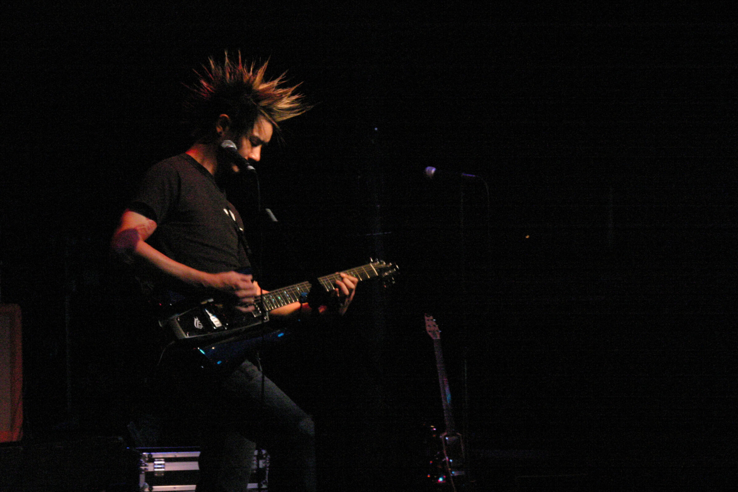 A guitarist with spiked hair plays an electric guitar into a microphone on a dark stage