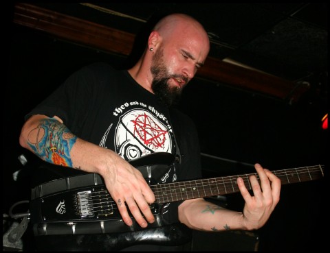A bald, bearded guitarist with tattooed arms plays a black RKS guitar on a dimly lit stage