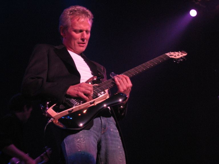 A guitarist in a black jacket and jeans plays a glossy black RKS guitar under purple stage lights