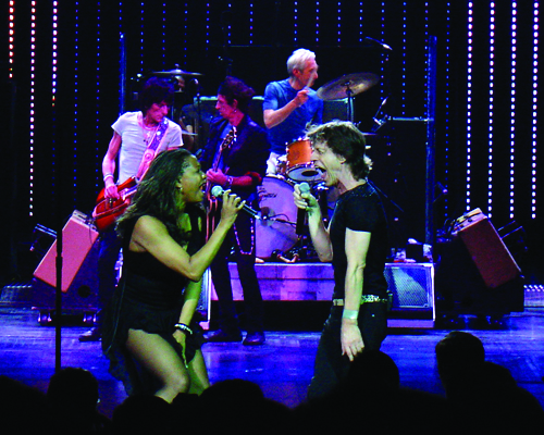 Two singers face each other at the front of a stage while two guitarists and a drummer play behind them
