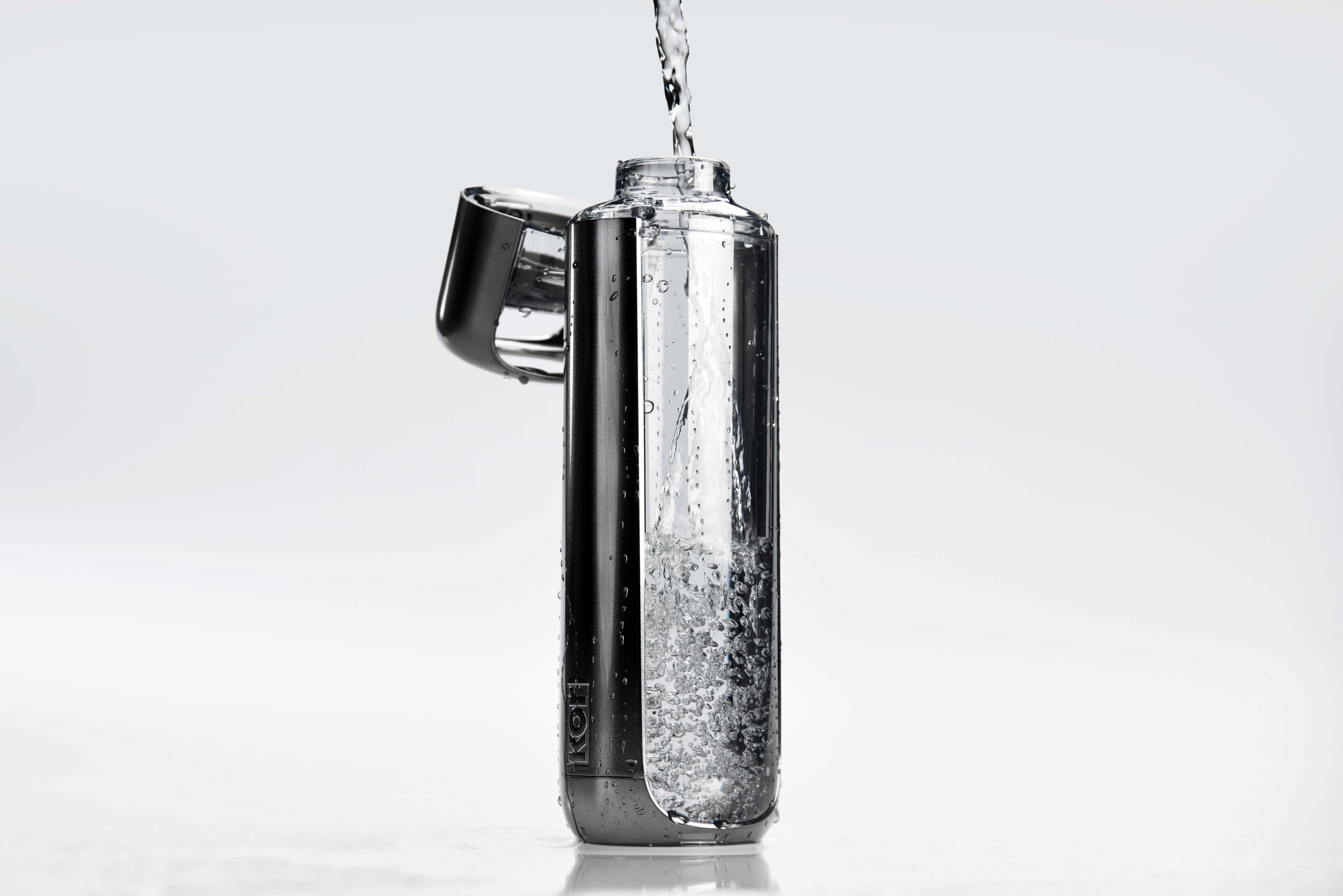 A clear, modern water bottle with a flip-top lid is being filled with water, shown…
