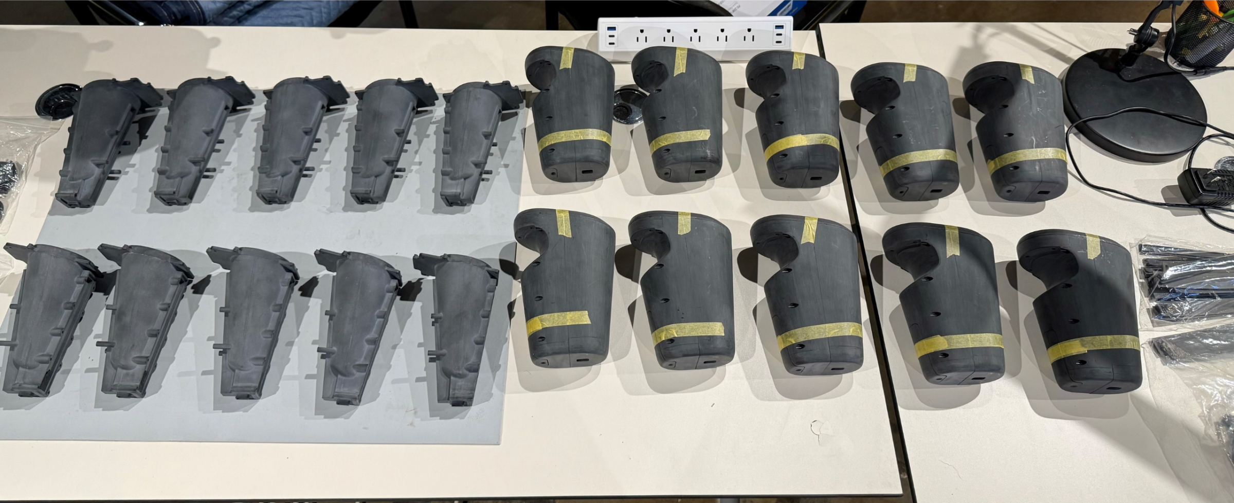 Two rows of black prototype housings and tapered inner parts, some marked with yellow tape, laid out on white tables
