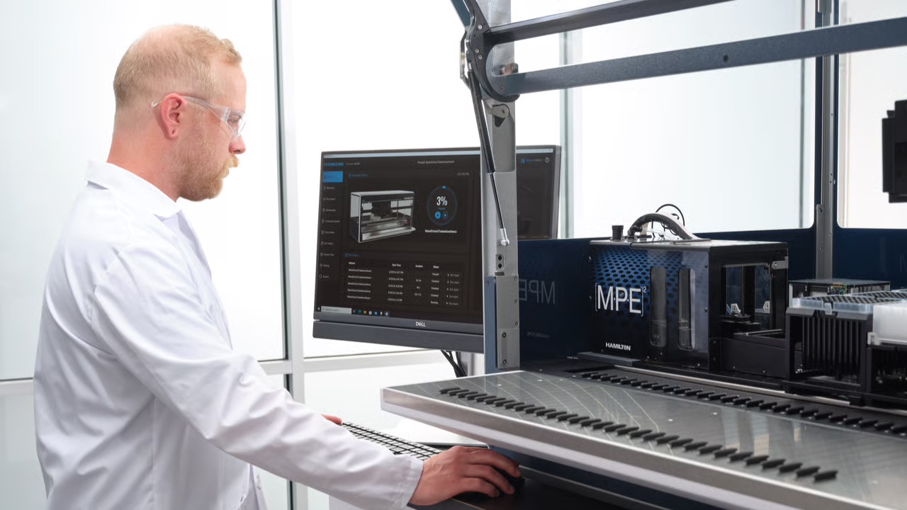 A person in a white lab coat and safety glasses operates laboratory equipment, using