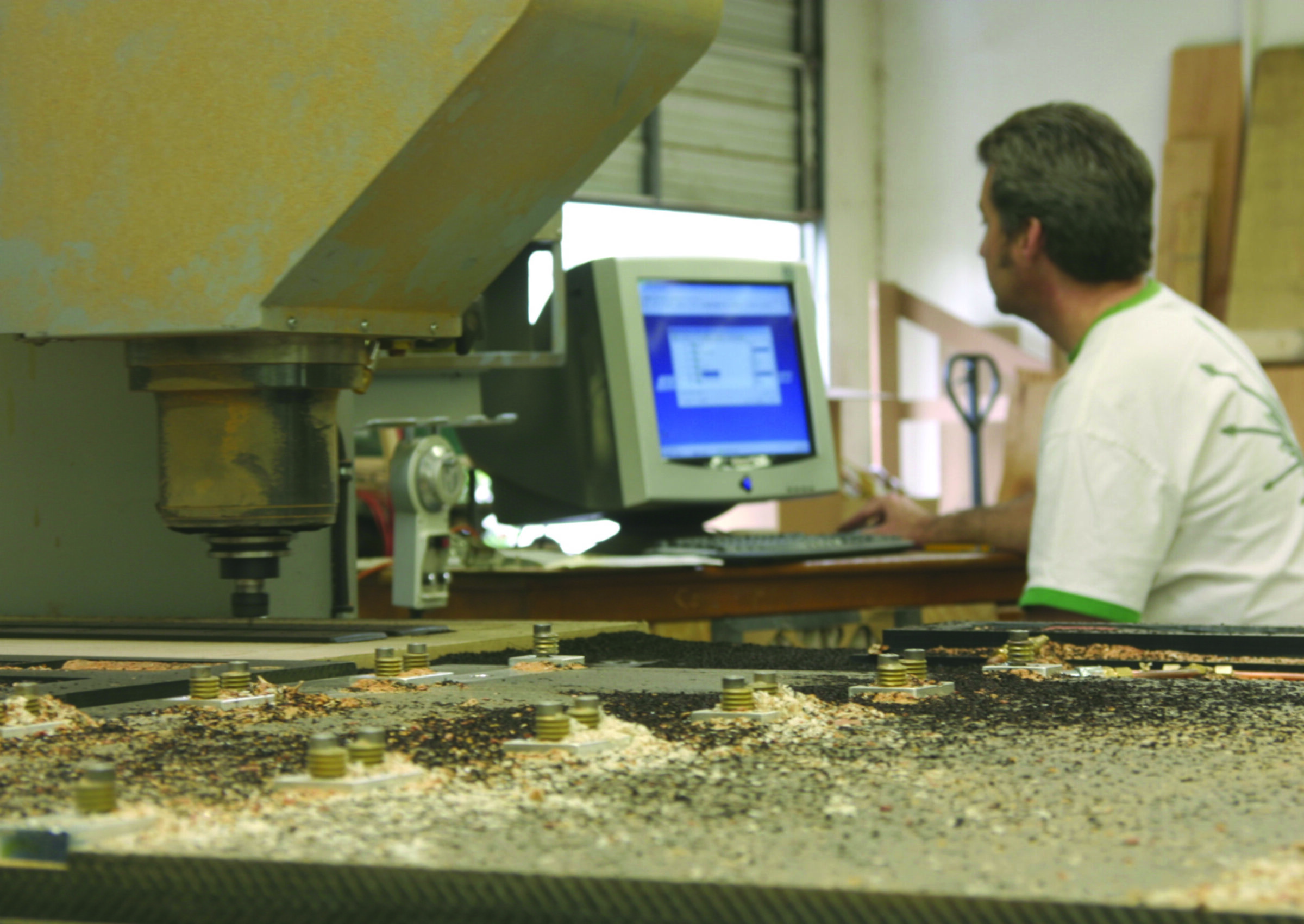 A man works at a computer beside a CNC router, its bed covered in wood shavings and hold-down clamps