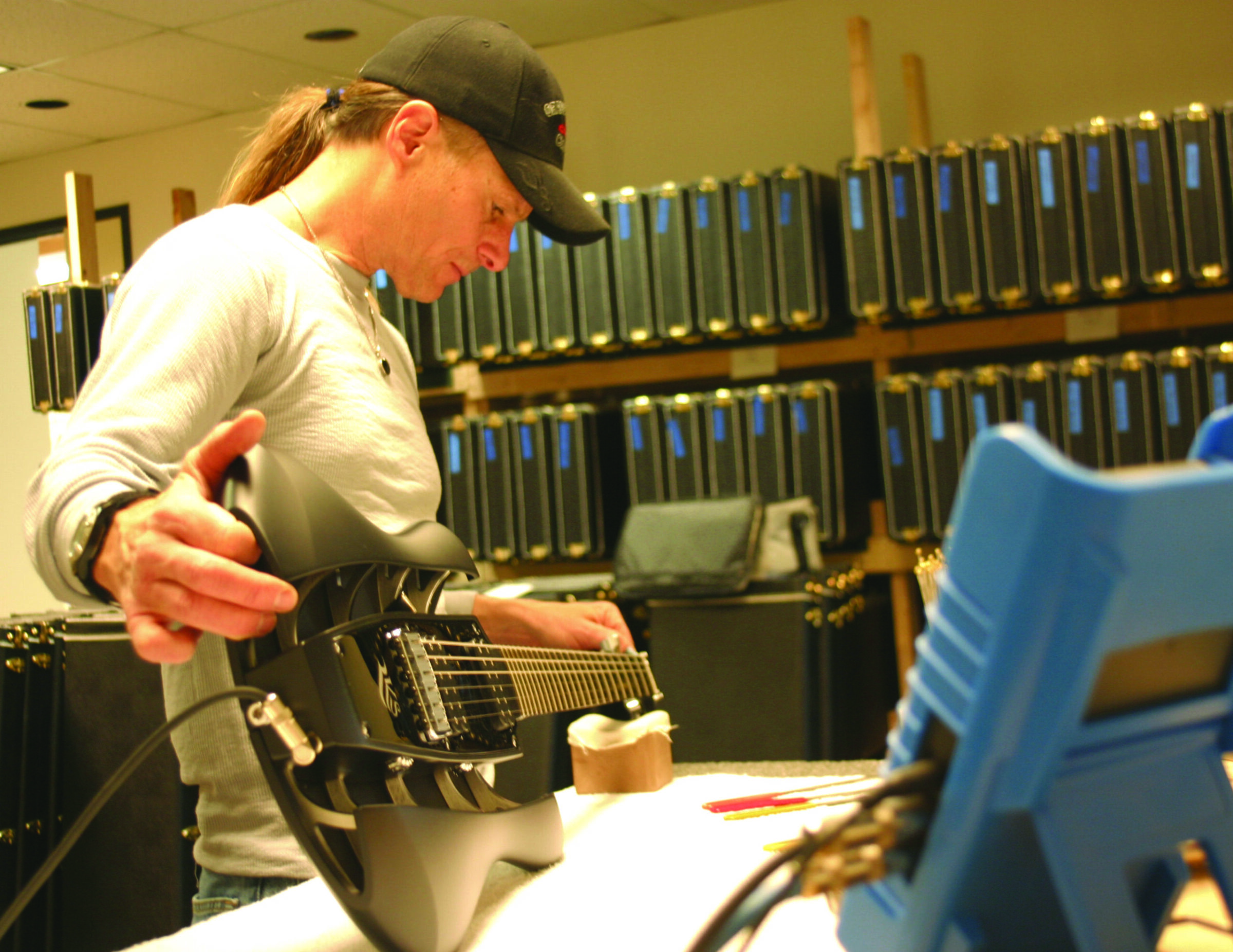 A man in a cap fits out a black RKS guitar at a workbench, with rows of guitar cases on shelves behind him