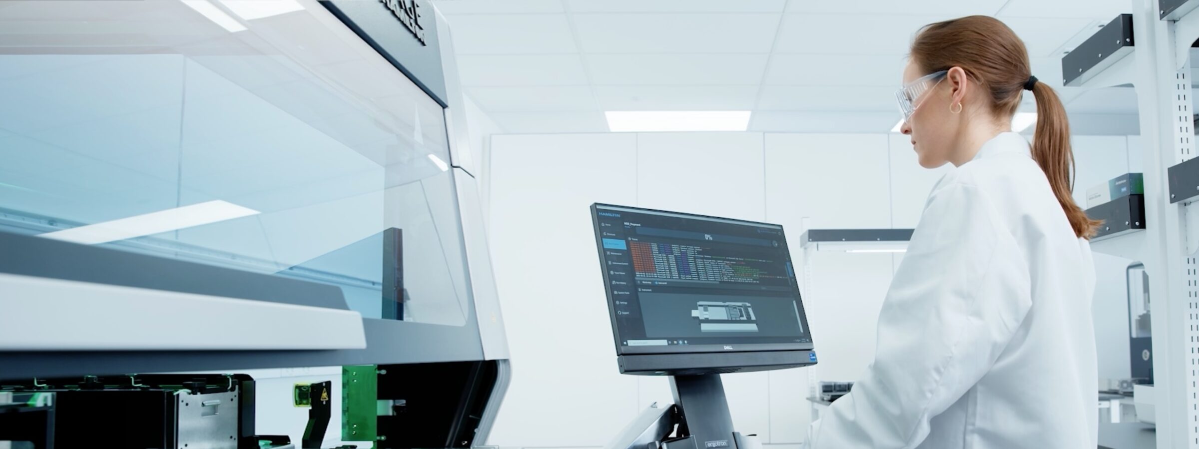 A scientist in a white lab coat and safety glasses works at a computer monitor displaying