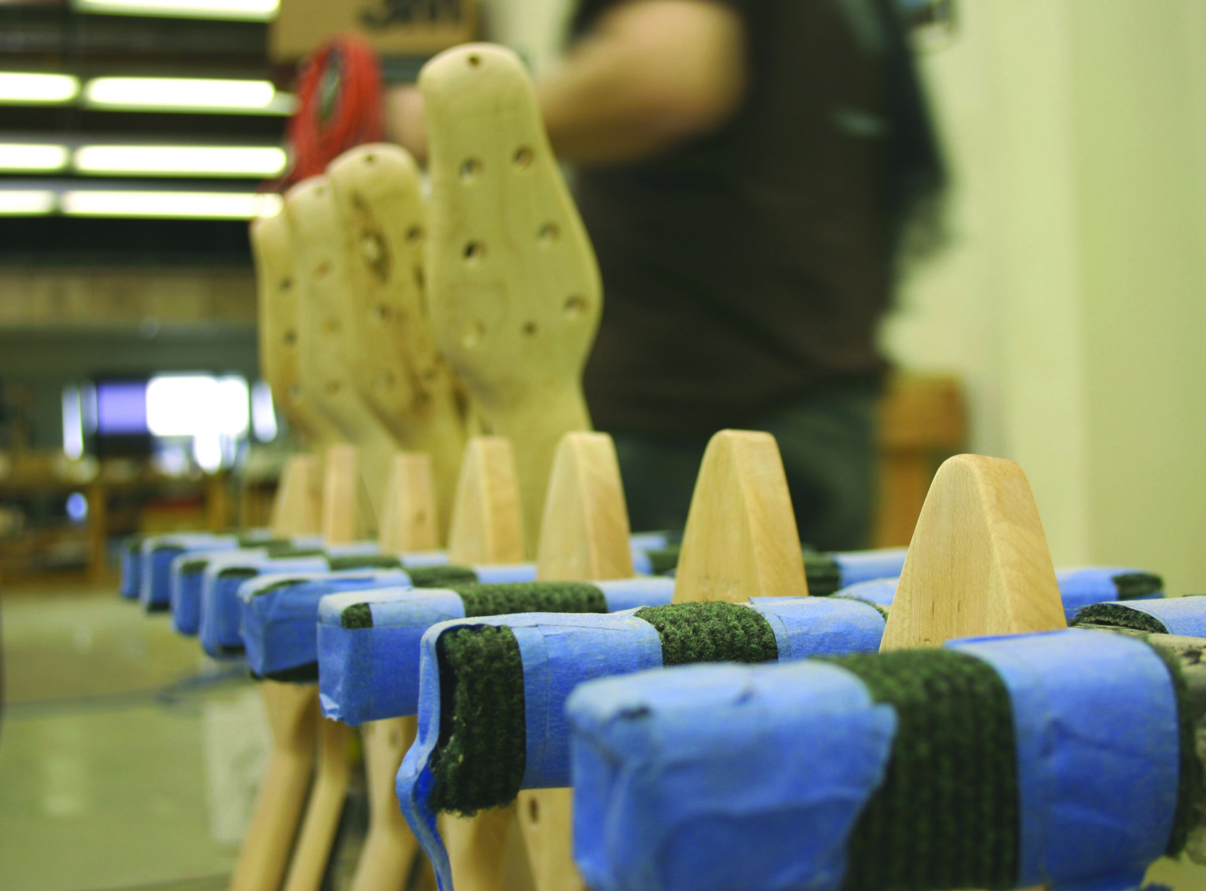 A row of unfinished wooden guitar necks with drilled headstocks, padded with blue tape and felt, in a workshop