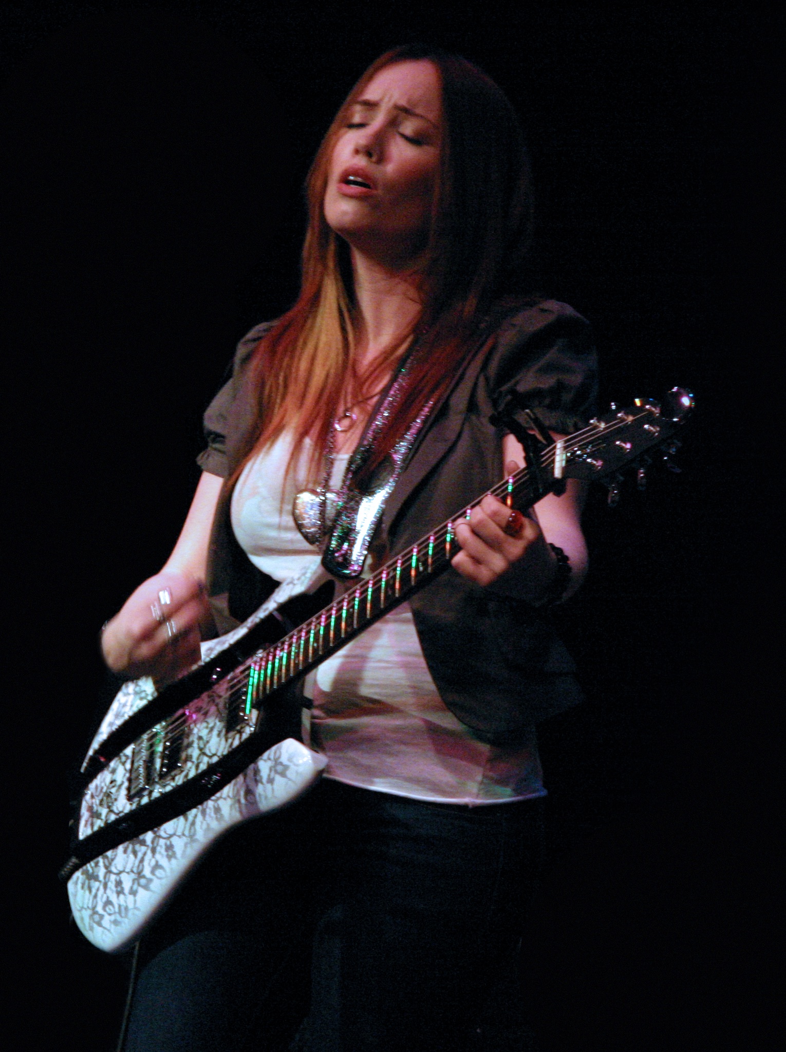 A woman with long hair plays a white lace-patterned electric guitar with lit fret markers on a dark stage