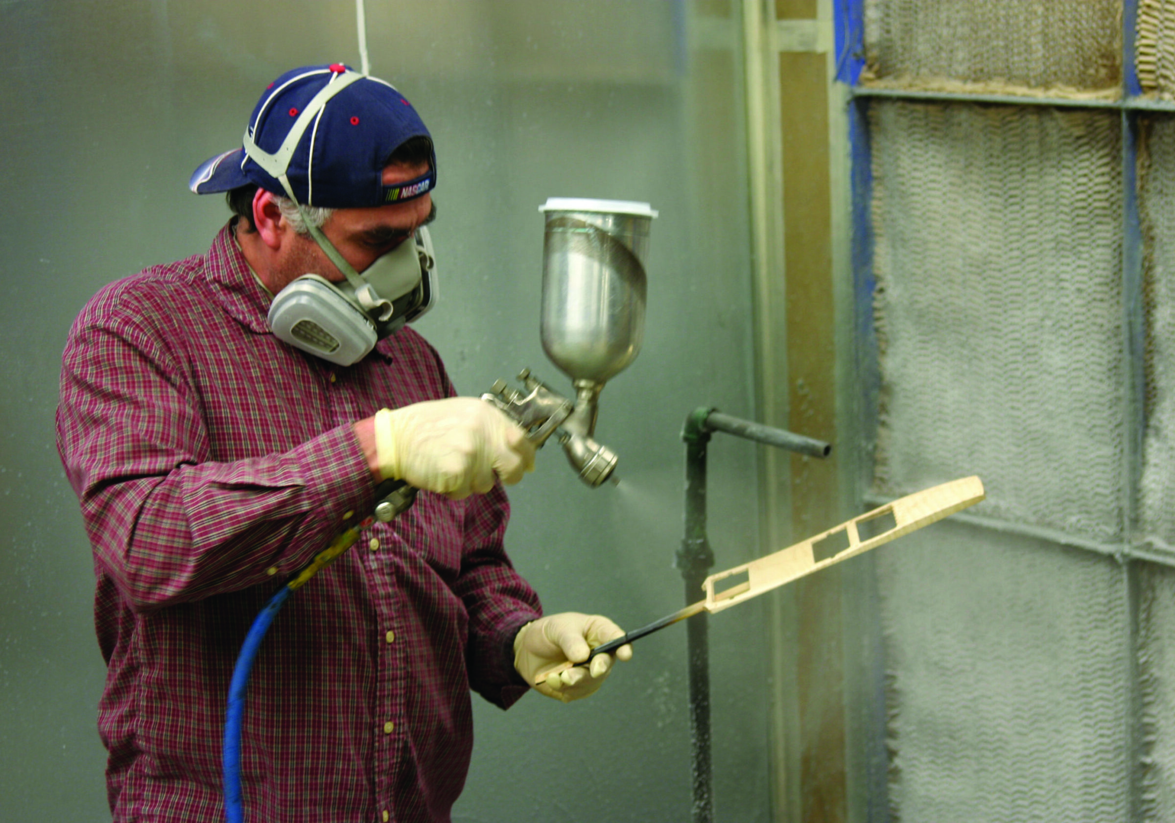 A man in a respirator and gloves spray-finishes a slotted wooden guitar part in a paint booth