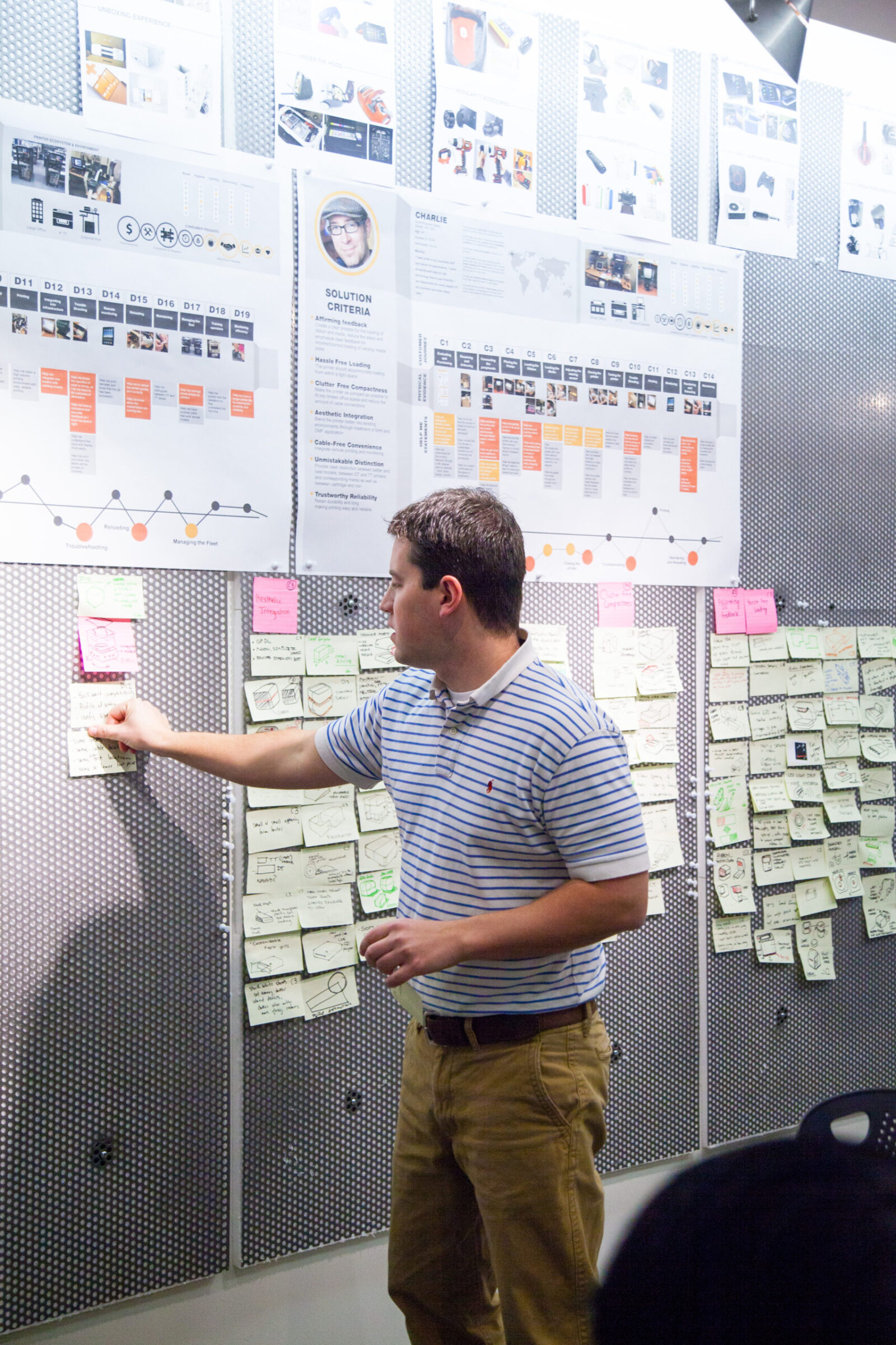 A man points to sticky notes on a wall below research posters with personas, timelines and solution criteria