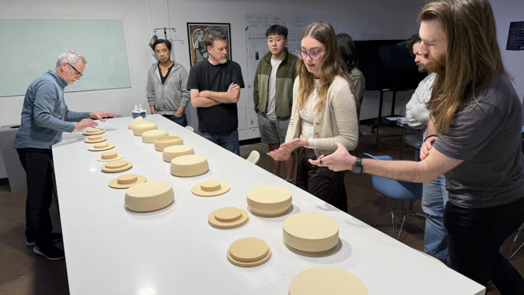 A group of people stand around a long white table with several round, beige objects…