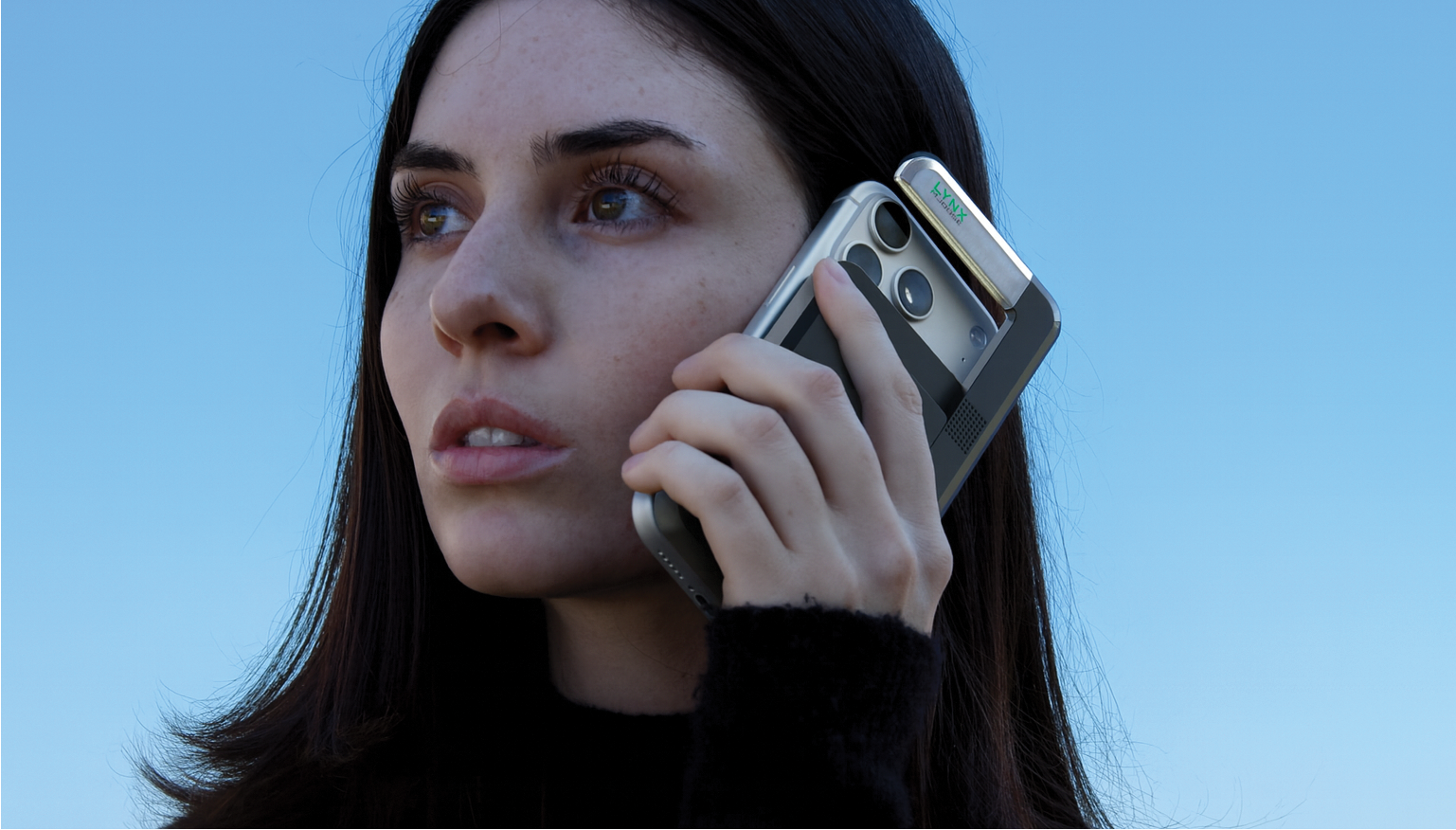 A woman with long dark hair holds a slim silver phone to her ear, looking thoughtful.