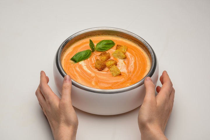 A person holds a bowl of creamy tomato soup topped with croutons and basil leaves…