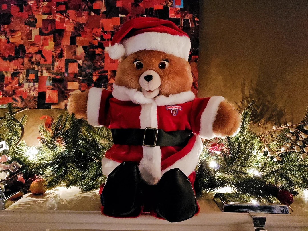 A Teddy Ruxpin dressed in a red and white Santa Claus outfit sits on a mantle decorated…