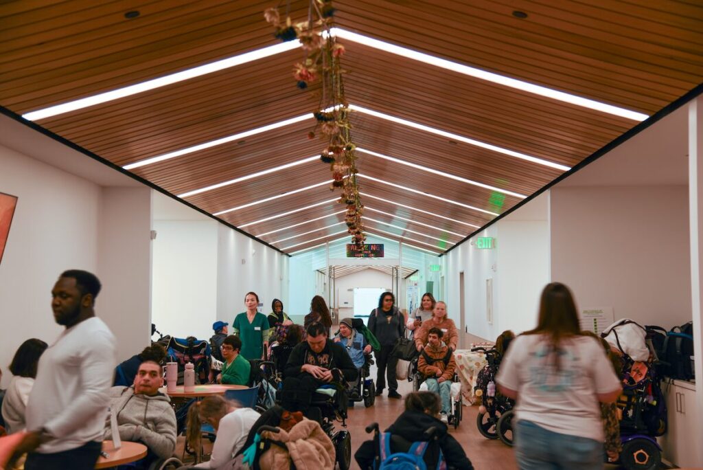A busy hallway is filled with people, some in wheelchairs and others standing or walking.
