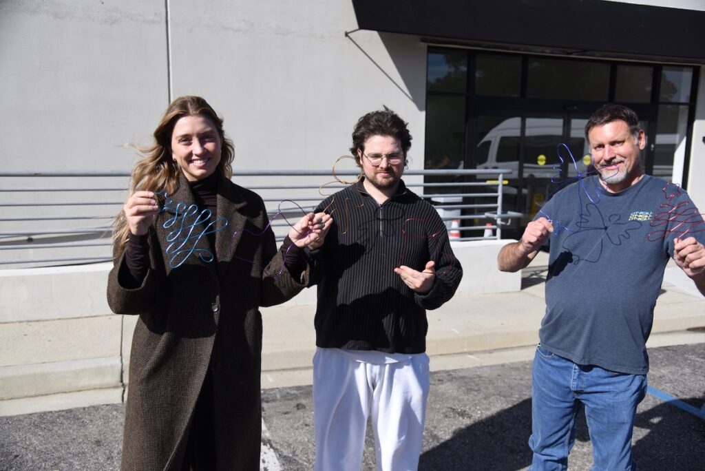 Three people stand outside on a sunny day, smiling and holding up colorful wire…