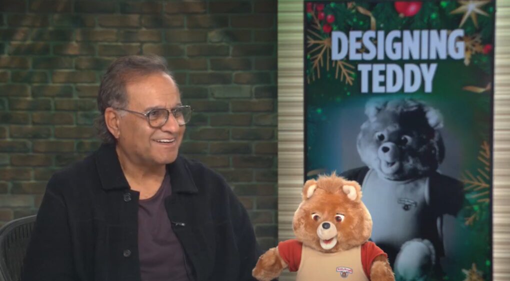 A smiling man sits next to a Teddy Ruxpin animatronic toy, with a poster behind…