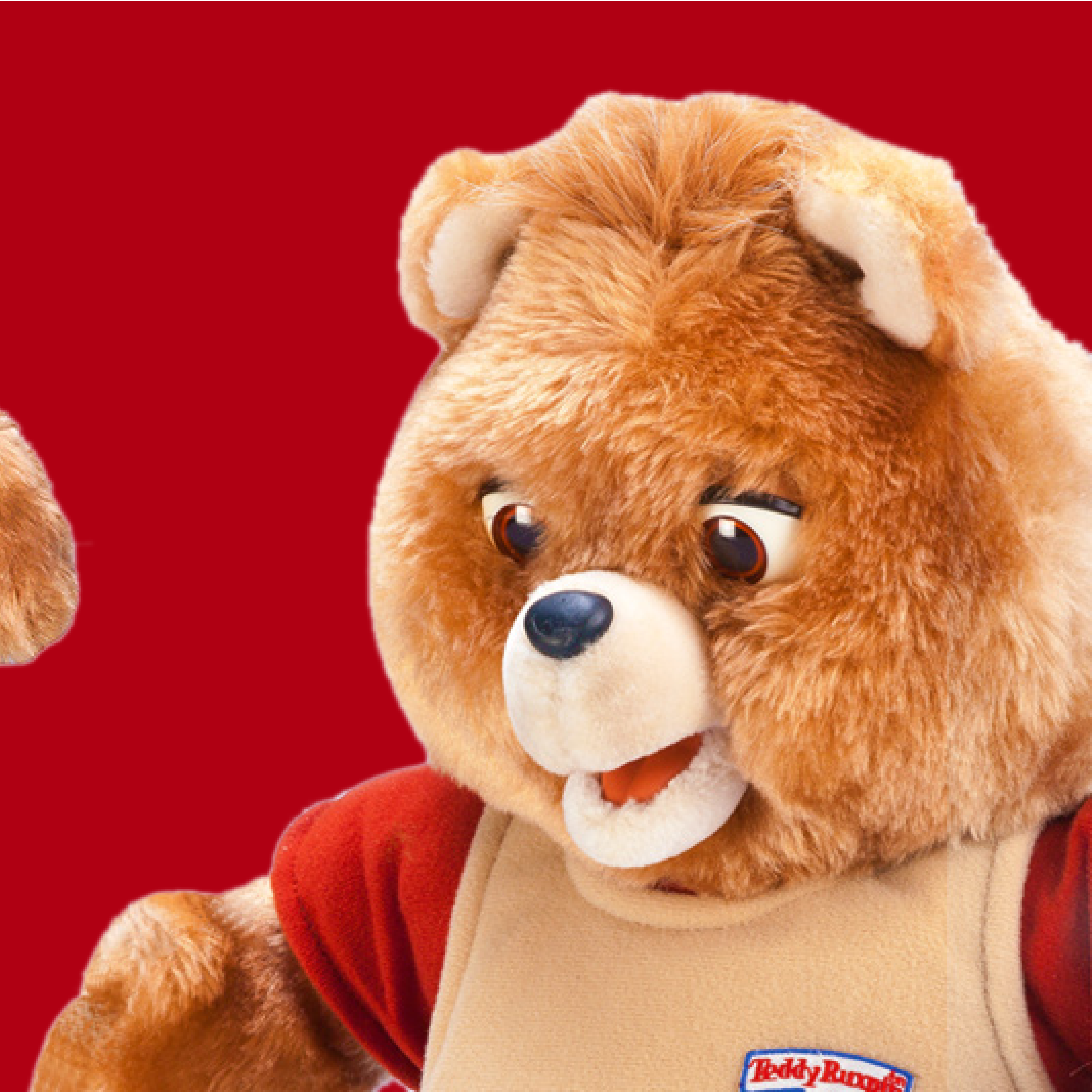 A Teddy Ruxpin bear with its mouth open and eyes glancing down, wearing its red and tan shirt, on a red background