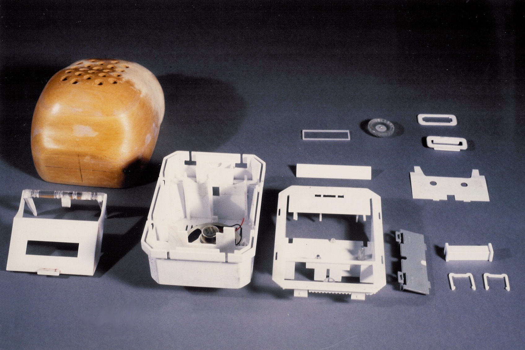 A wooden form model beside disassembled white plastic housing parts and a small speaker, laid out on a gray surface