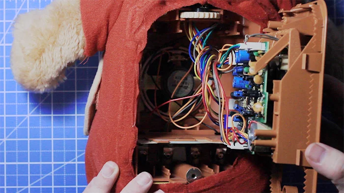 Hands open the back of a Teddy Ruxpin bear, exposing wiring, a speaker and a circuit board inside its plastic frame