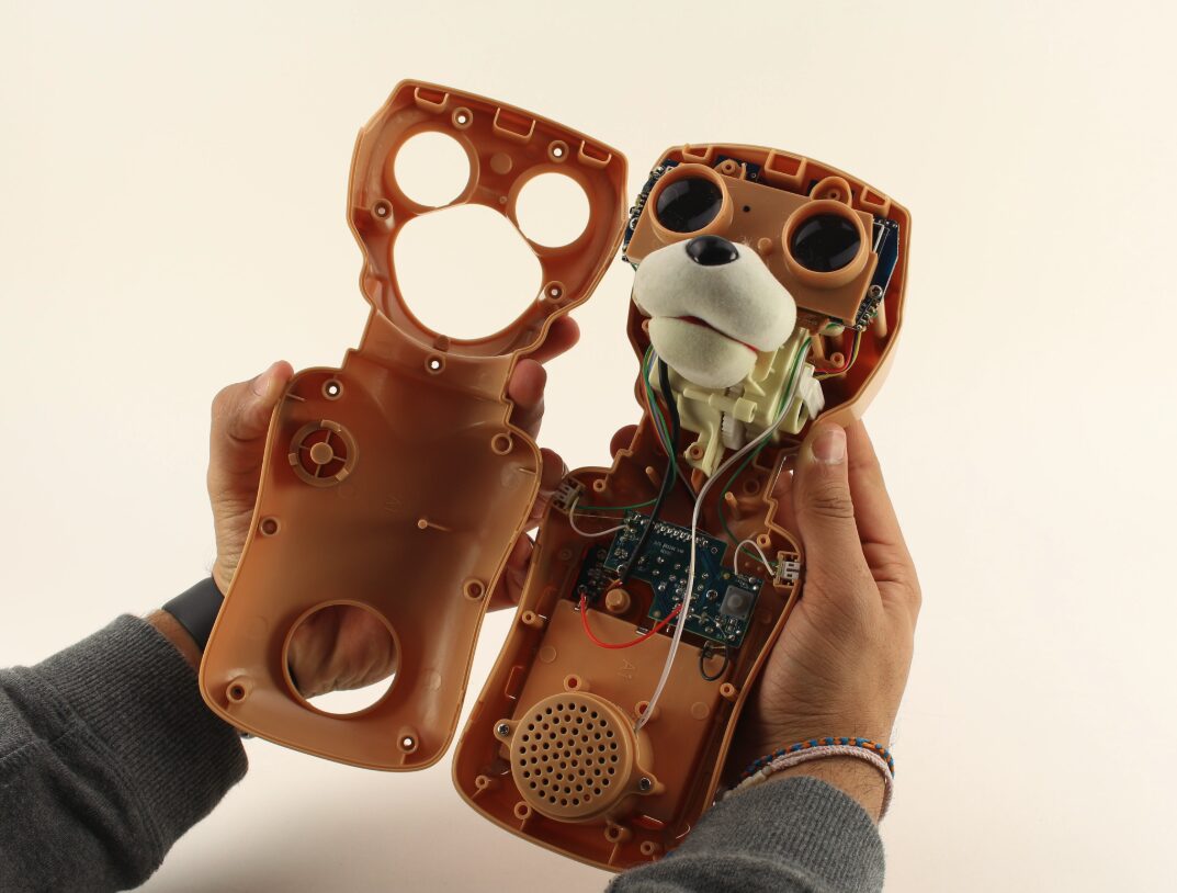 Hands hold open the plastic body shell of a Teddy Ruxpin bear, showing its eyes, snout, circuit board and speaker