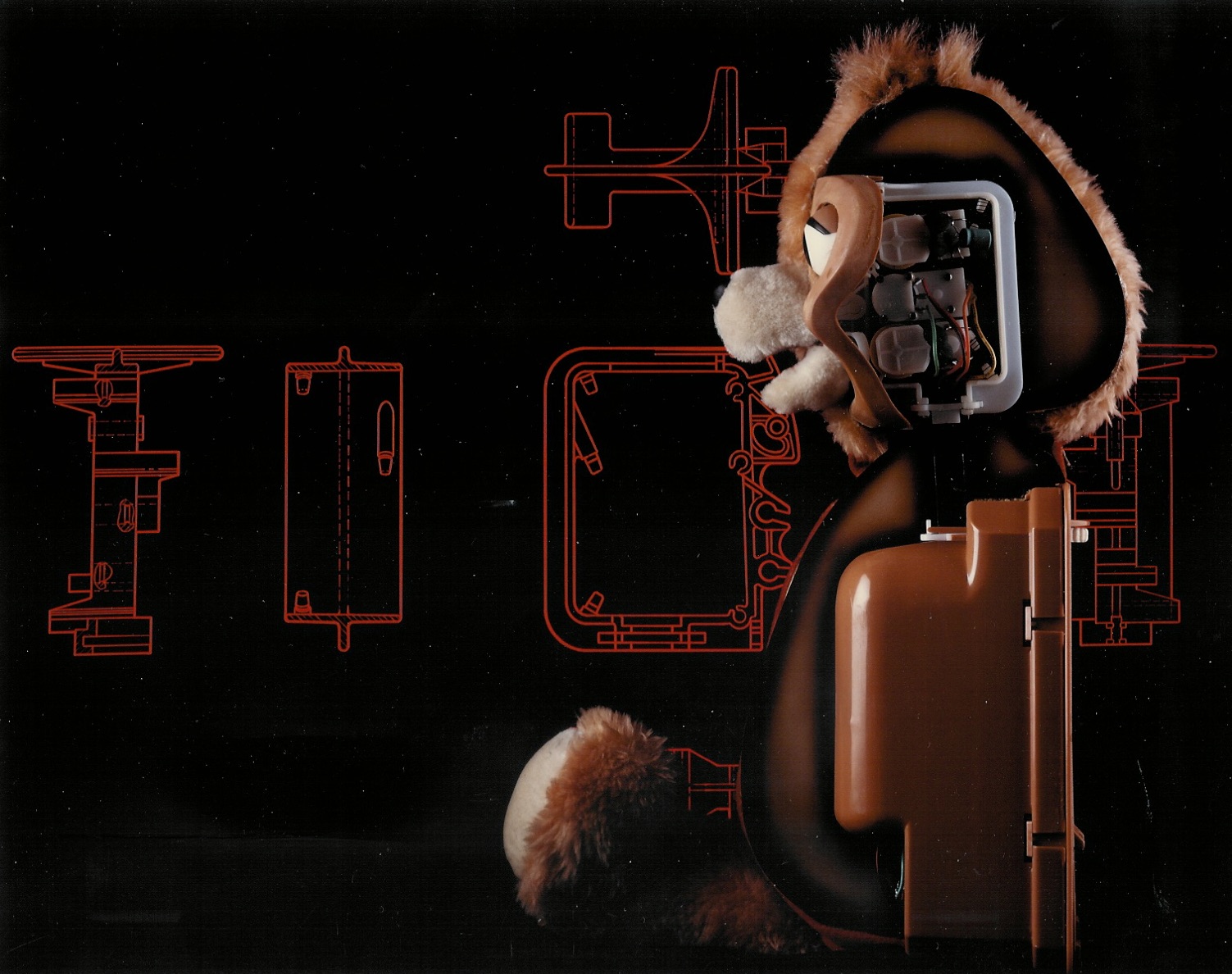 A partly opened Teddy Ruxpin bear showing its internal mechanism and plastic housing, beside red technical drawings
