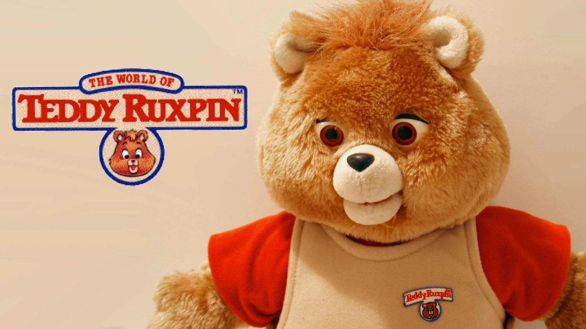 A plush Teddy Ruxpin bear with tan fur, a red shirt, and a beige vest stands beside…