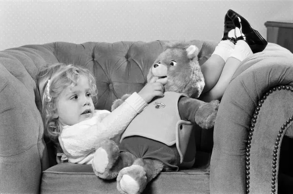 Black and white photo of a young girl lounging on a tufted sofa with a Teddy Ruxpin bear