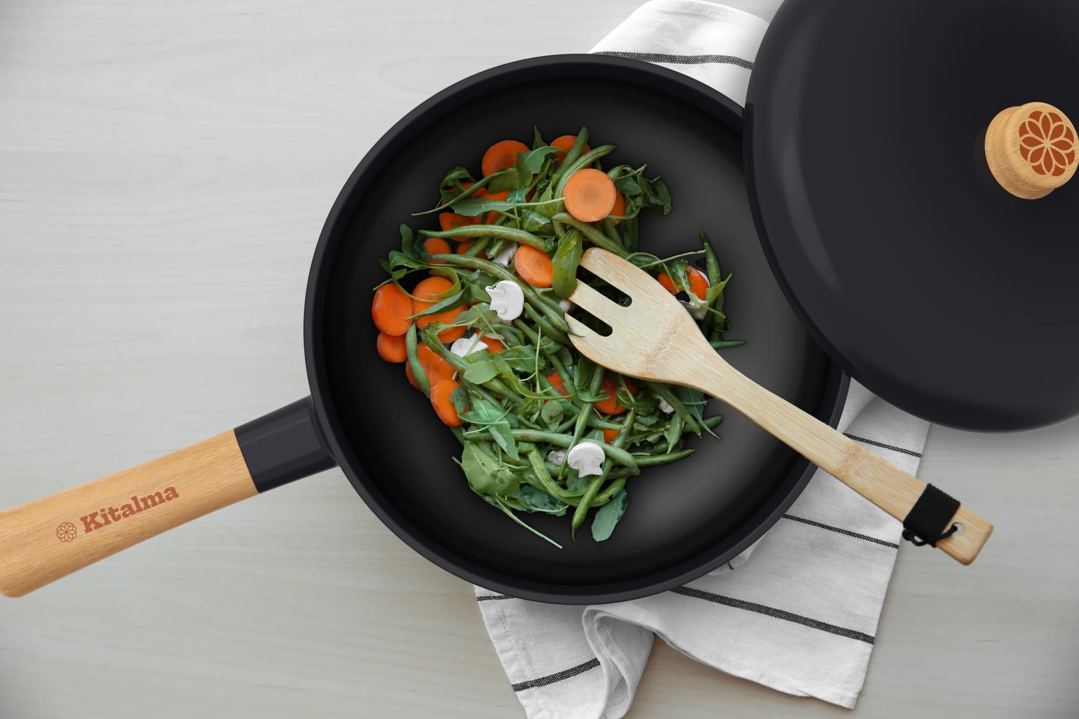 A Kitalma wok of greens and carrots seen from above, with its lid and a wooden spatula.