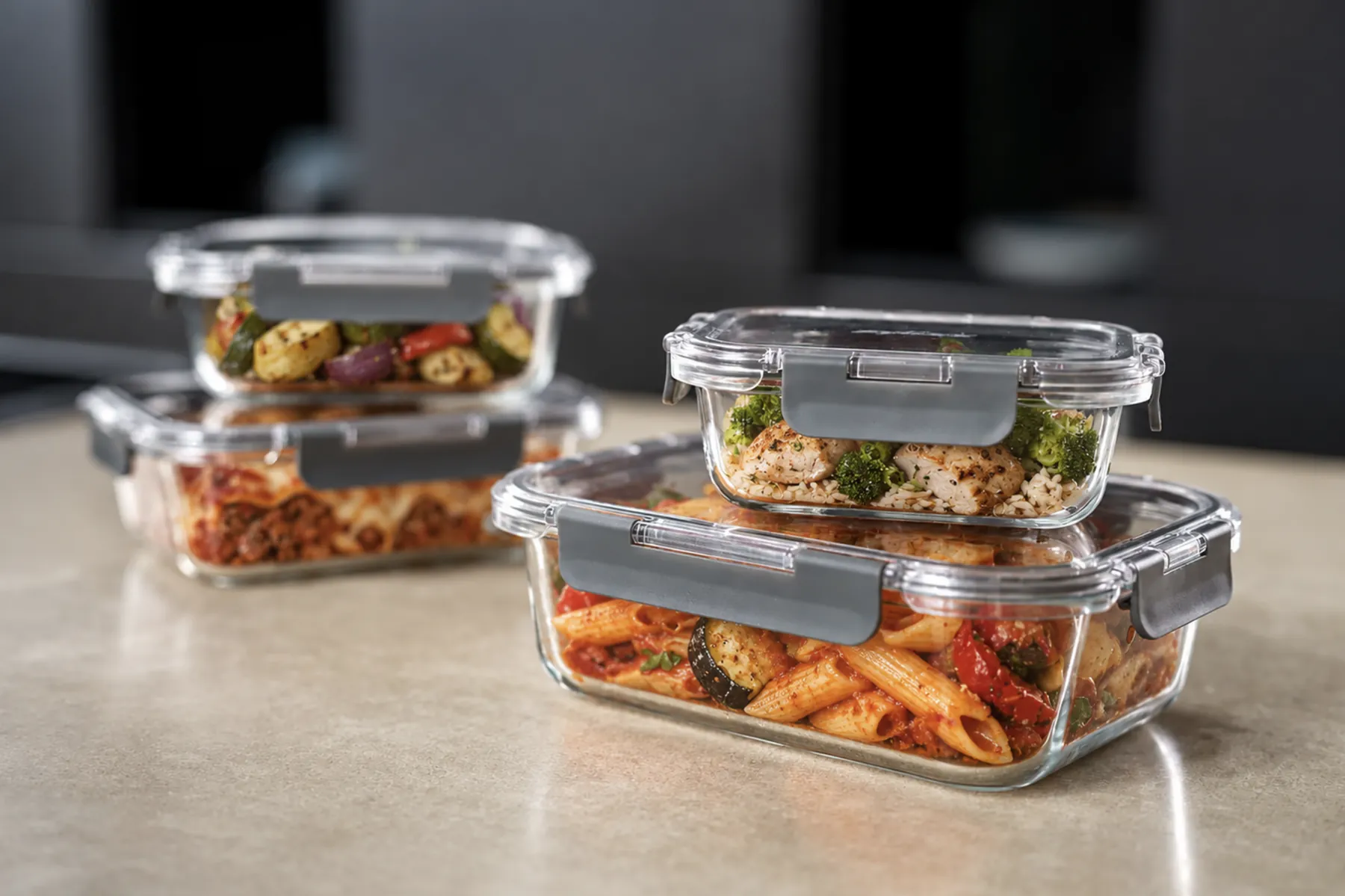Four Zyliss glass food storage containers with grey latching lids, filled with penne, lasagna, chicken and rice, and roasted vegetables, stacked on a kitchen countertop.
