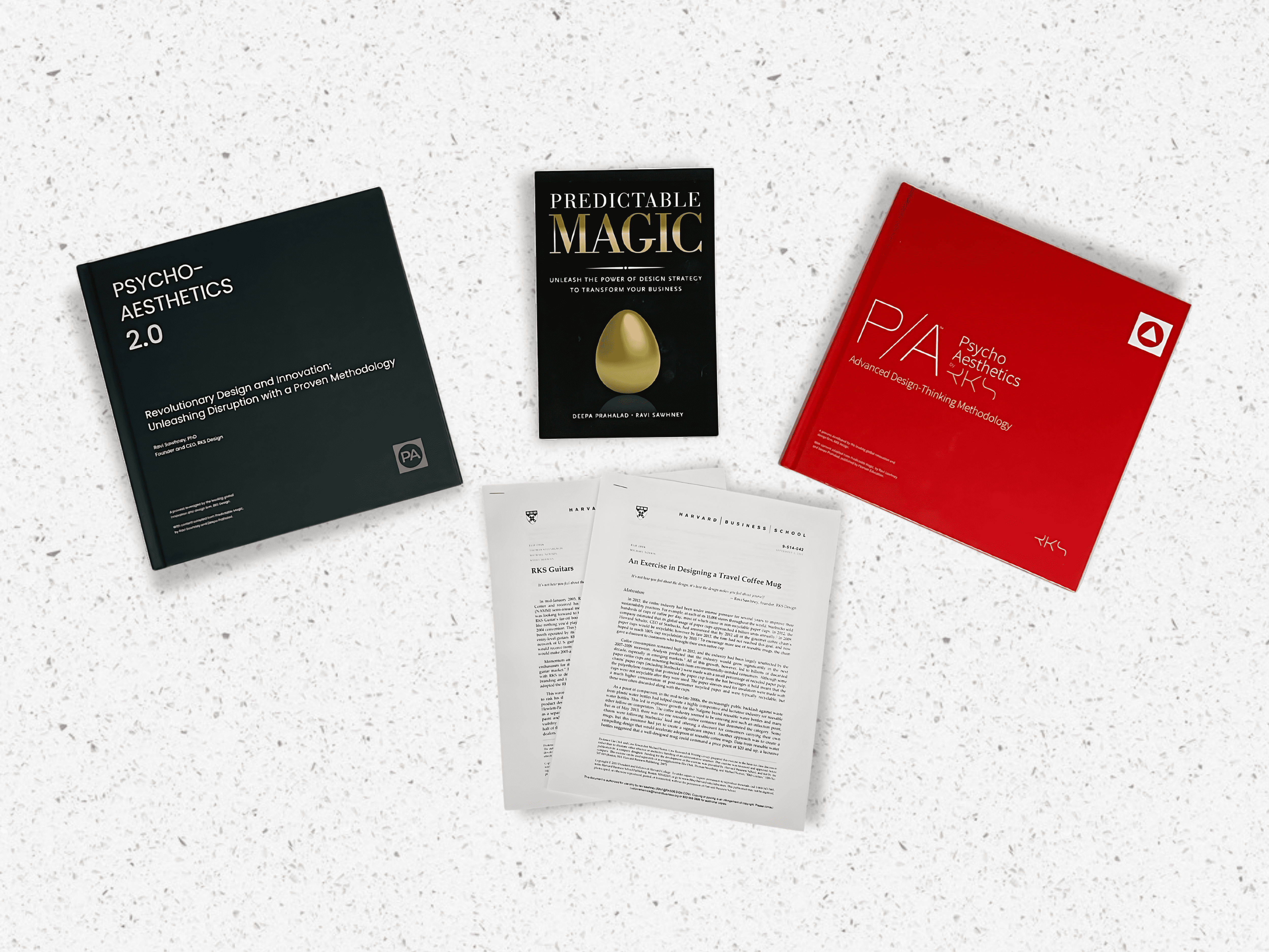 A collection of design and innovation books and documents on a white speckled surface. The items include "Psycho-Aesthetics 2.0" with a black cover, "Predictable Magic" with a black cover and a golden egg on it, "Psycho Aesthetics" with a red cover, and two documents with text, including one titled "An Exercise in Designing a Travel Coffee Mug
