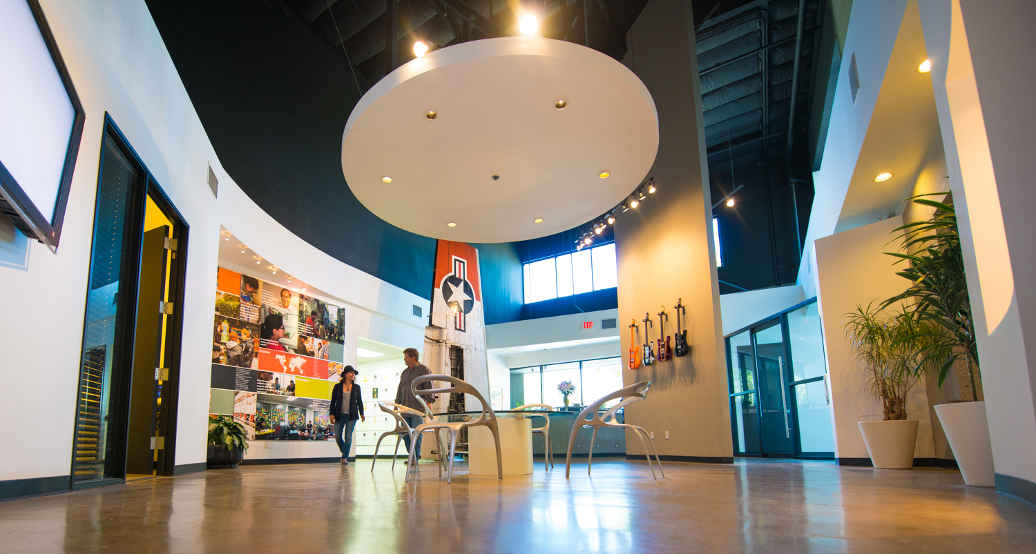 Modern office lobby with a circular ceiling feature, polished concrete floors, and sleek metallic chairs around a small table. A large airplane tail decorates the center, and colorful wall graphics display images of people and global initiatives. On one side, electric guitars are mounted on the wall, while on the other, large windows let in natural light, enhancing the open and welcoming atmosphere.