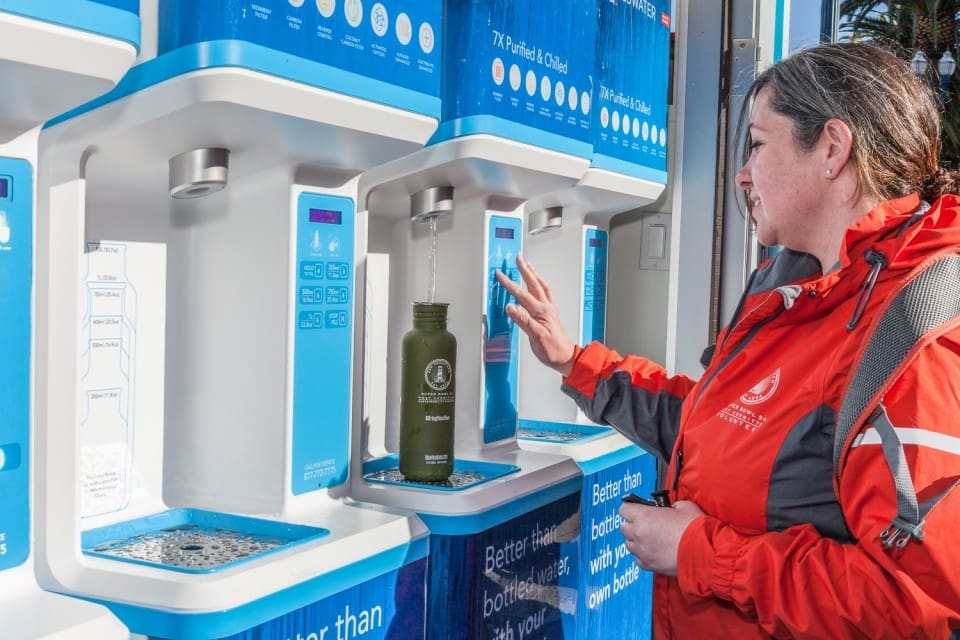 A woman refills a reusable water bottle at a public hydration station, pressing a button as filtered water dispenses into her bottle. She is wearing a red jacket and standing outdoors.