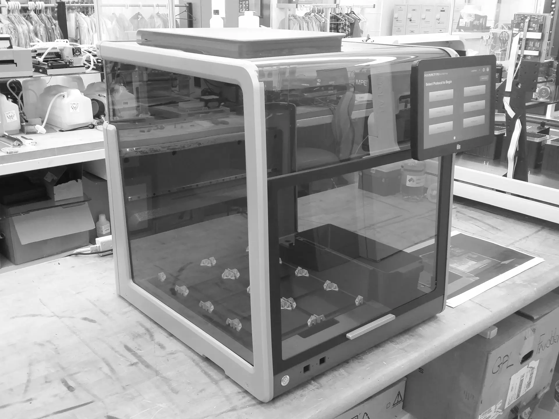 A large 3D printer with a touchscreen interface sits on a workbench in a workshop, surrounded by tools, equipment, and shelves holding various supplies.