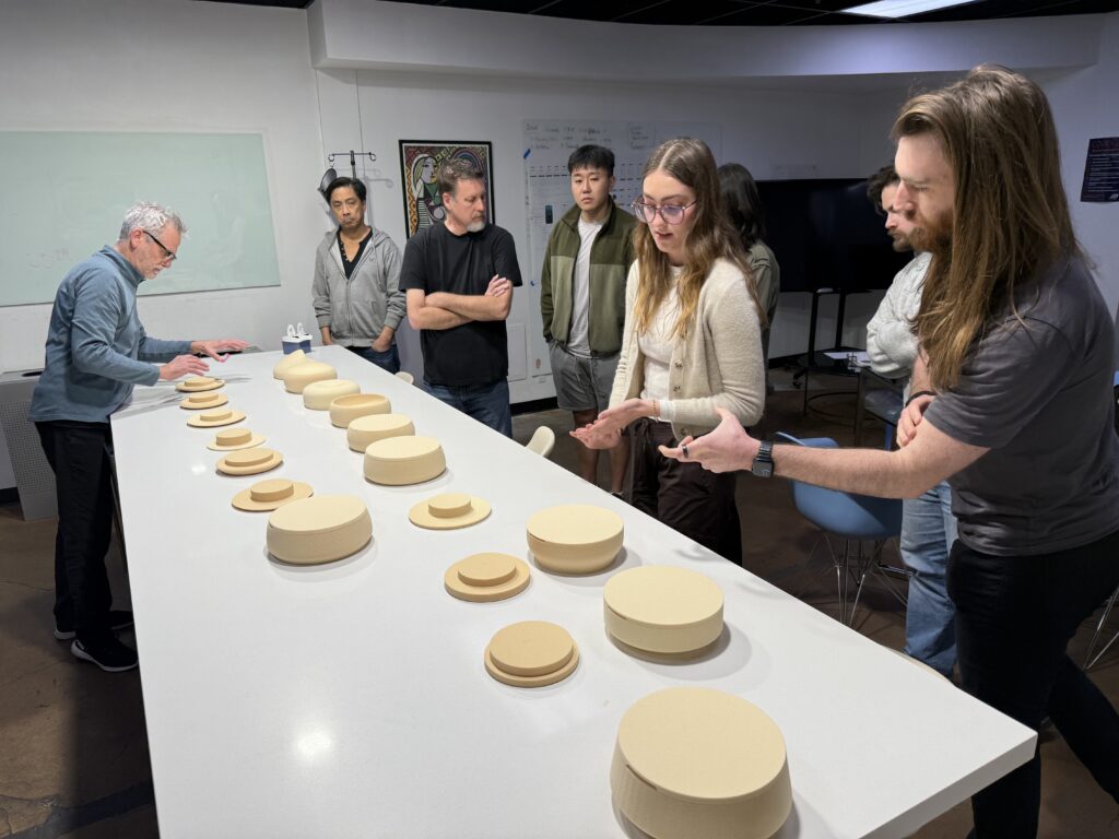 A group of people stand around a white table with variously sized beige circular objects. One person gestures towards the objects while others watch. A person at the end of the table appears to be arranging the objects.