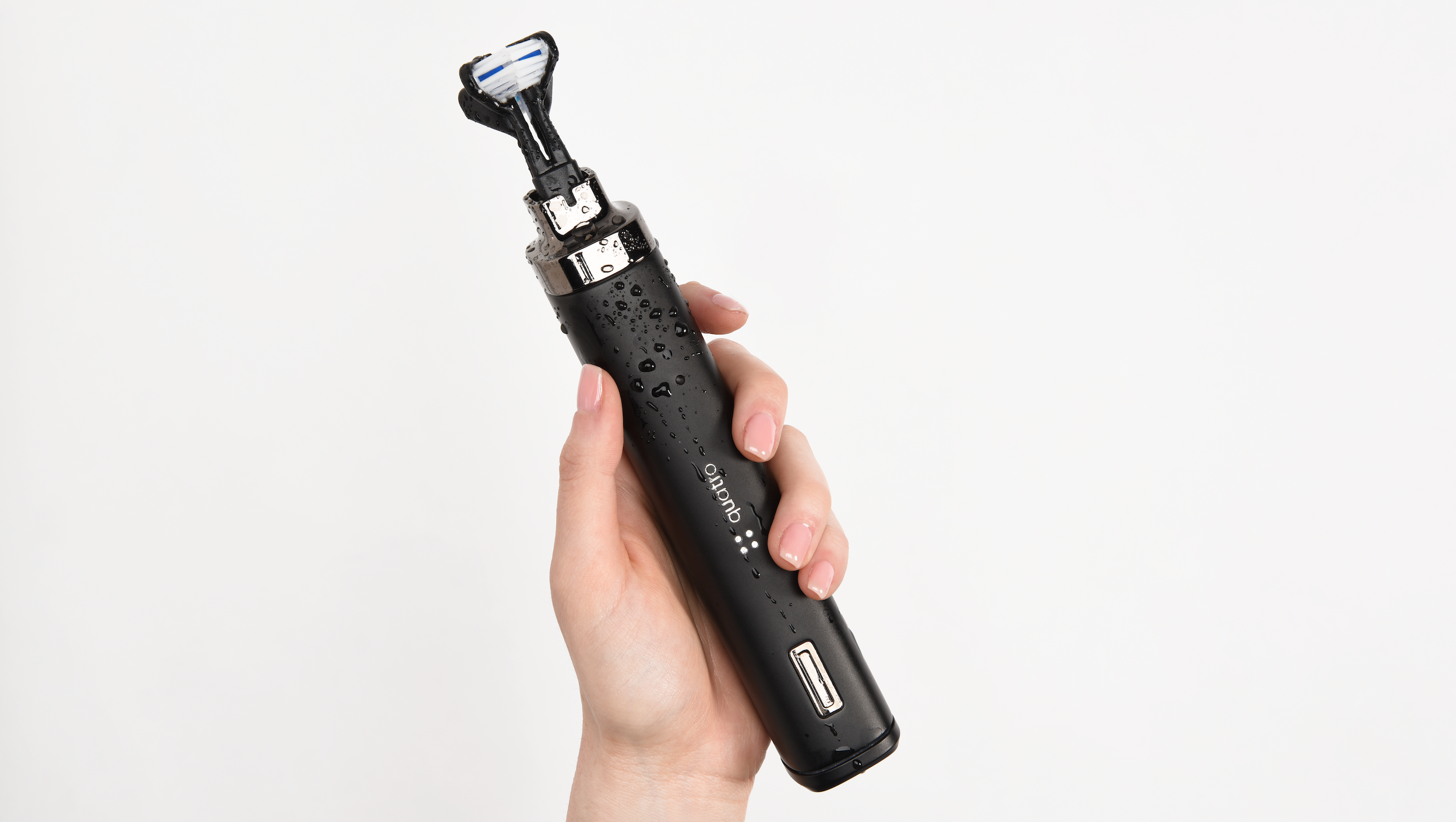 Hand holding the Quatro smart toothbrush with visible water droplets on a matte black handle, showing ergonomic design and dual-sided brush head