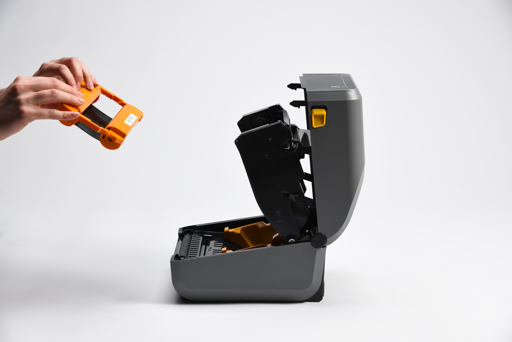 Hands placing orange ribbon cartridge above open Zebra printer with lid raised