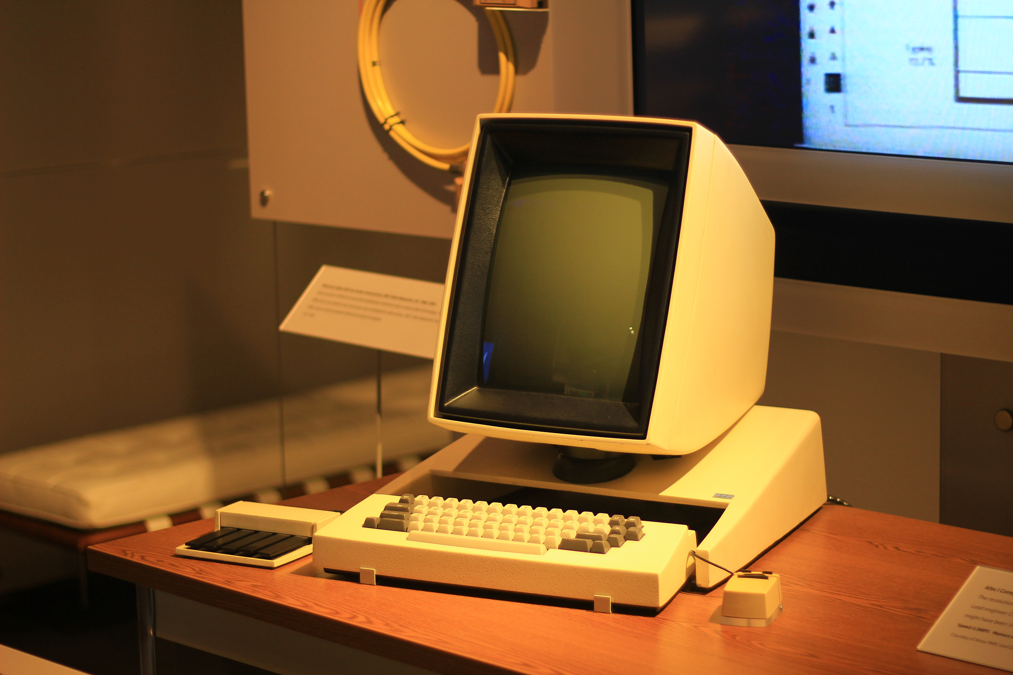 A vintage computer with a built-in monitor, keyboard, mouse, and keypad sits on a wooden desk in a museum exhibit. The screen is off and there are informational plaques nearby.