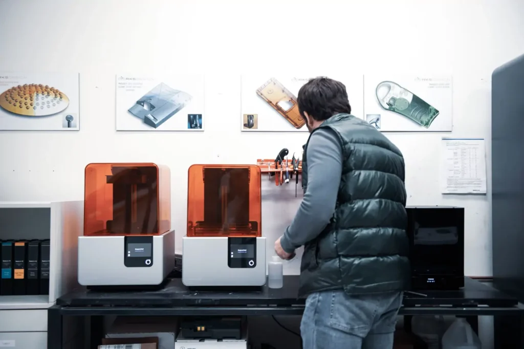 Engineer working with dual 3D printers in lab surrounded by concept printouts and shelving
