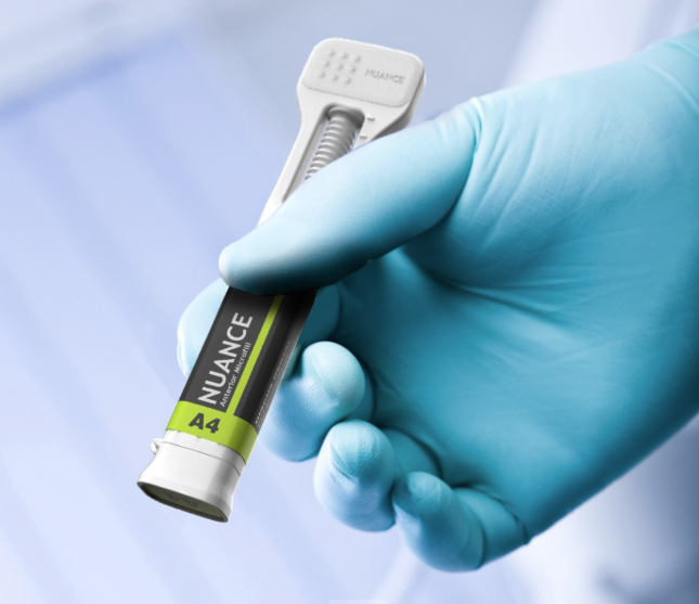 A gloved hand holds a dental composite syringe labeled NUANCE A4 against a blurred, light blue background, highlighting precision and quality for the medical market by AAA Medical in a clinical or dental setting.