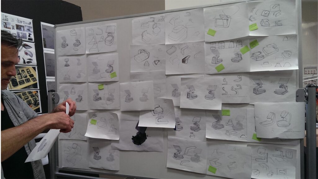 A man stands by a whiteboard covered with sheets of paper, each featuring sketches of various life science gadget or device designs. Some papers have green sticky notes attached. The man is holding additional papers.