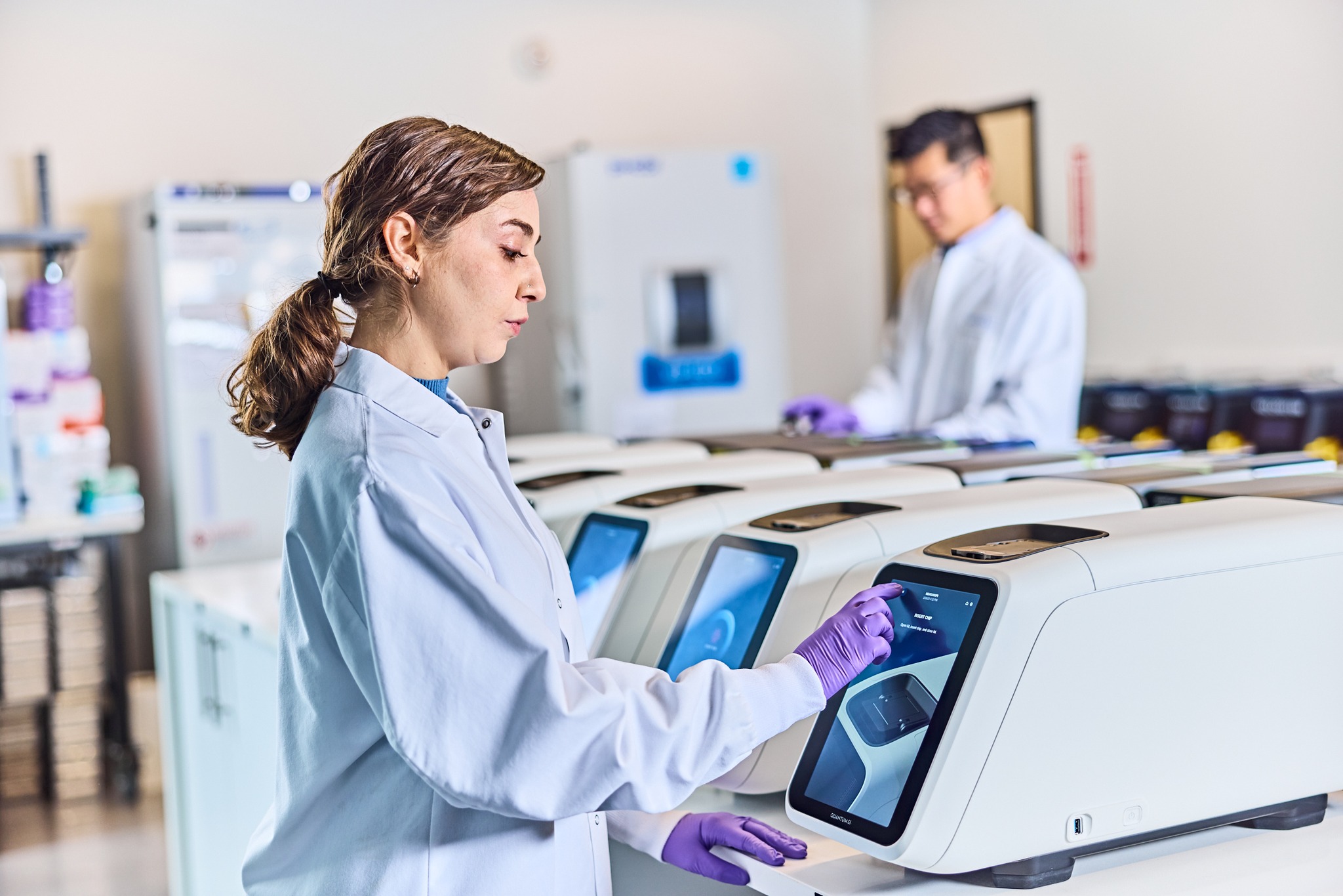 A woman in a lab coat and purple gloves uses a touchscreen on a laboratory instrument, with a man working in the background in a bright, modern lab setting.
