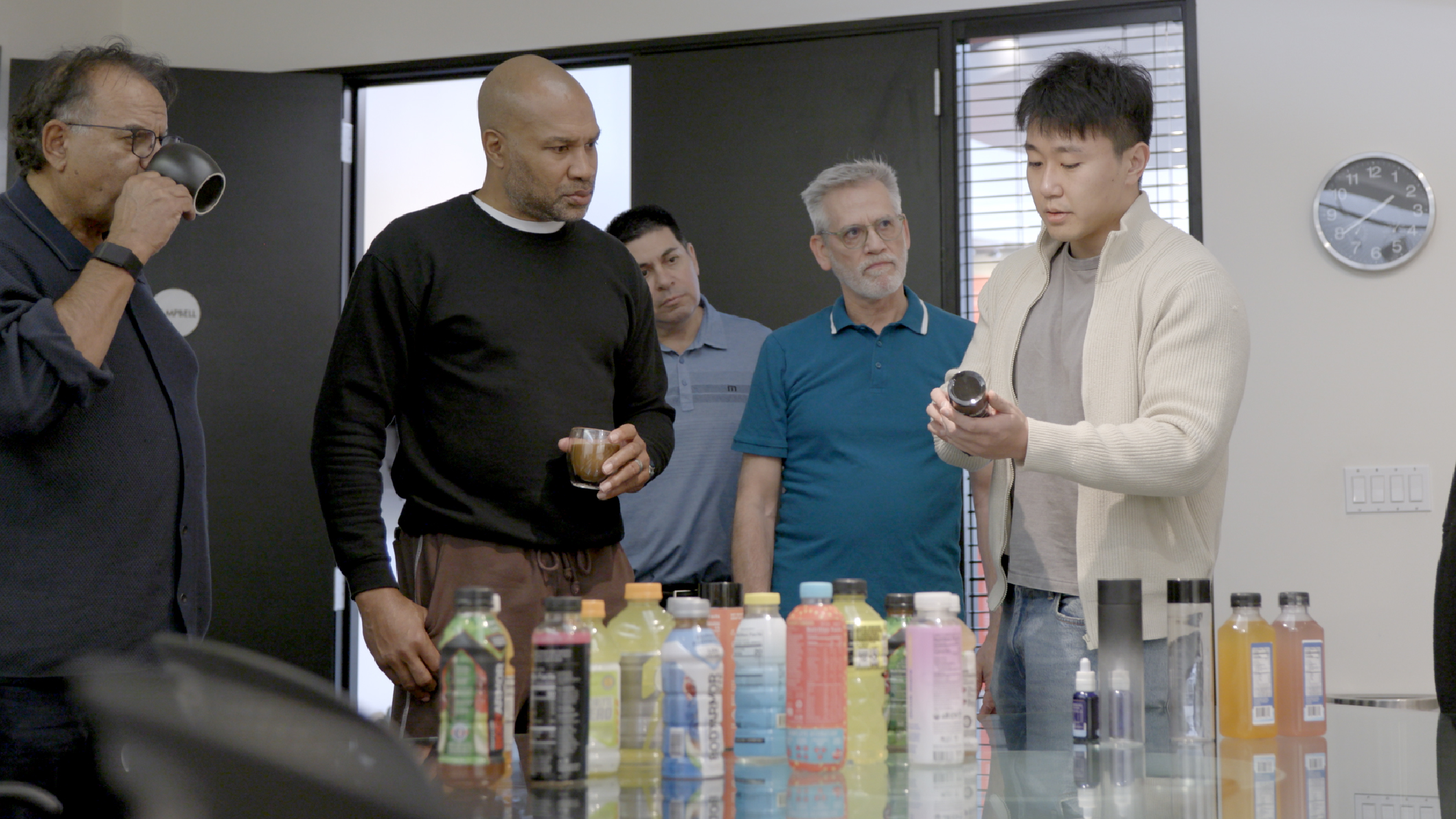 Five men stand around a table with various colorful beverage bottles. One man in a white cardigan is showing a bottle to the group while others watch or hold drinks. The setting appears to be an office or meeting room.