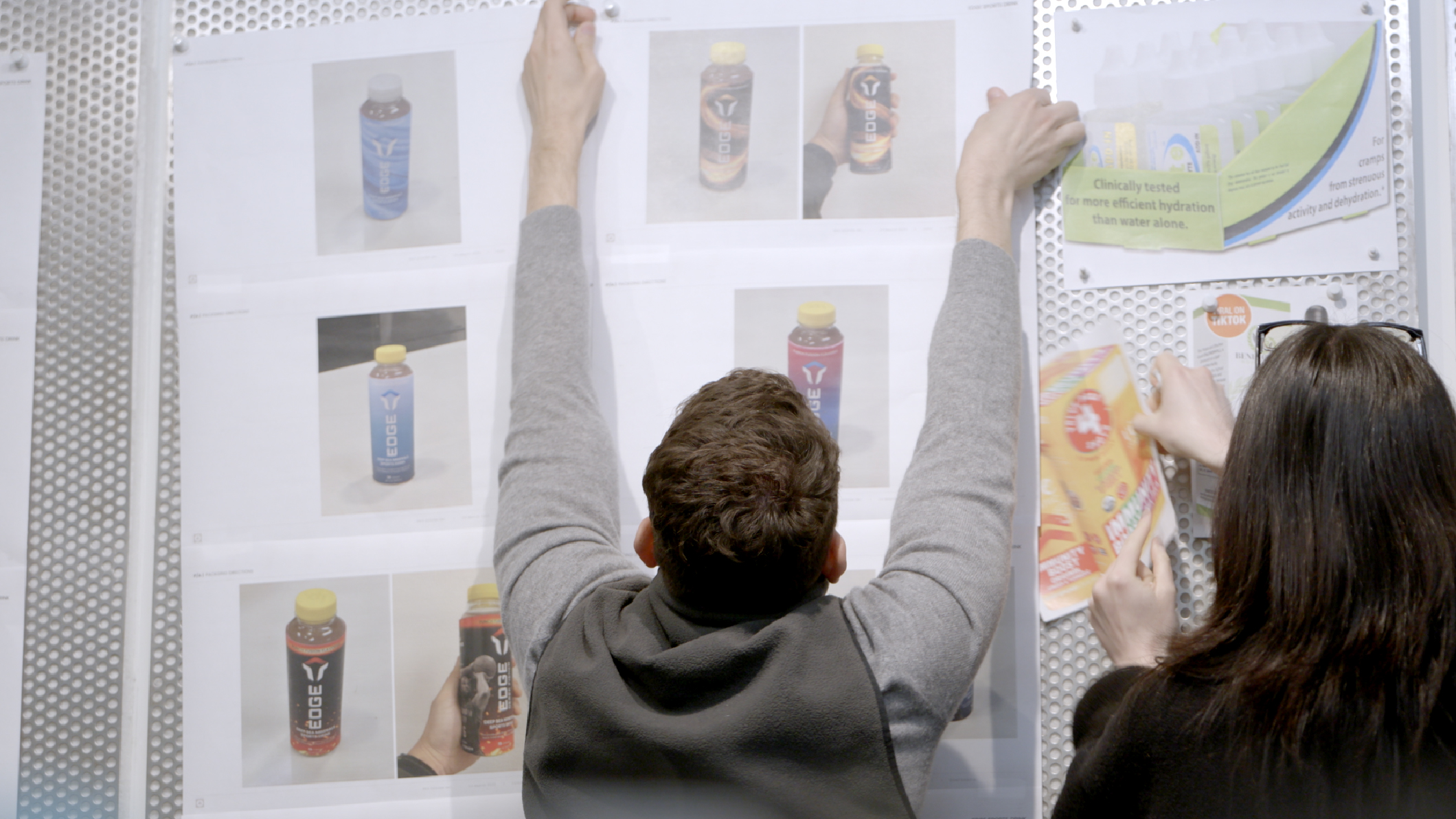 A person pins printed images of colorful beverage bottles on a wall, while another person holds papers nearby. Both are focused on arranging the images, suggesting a design or planning session.