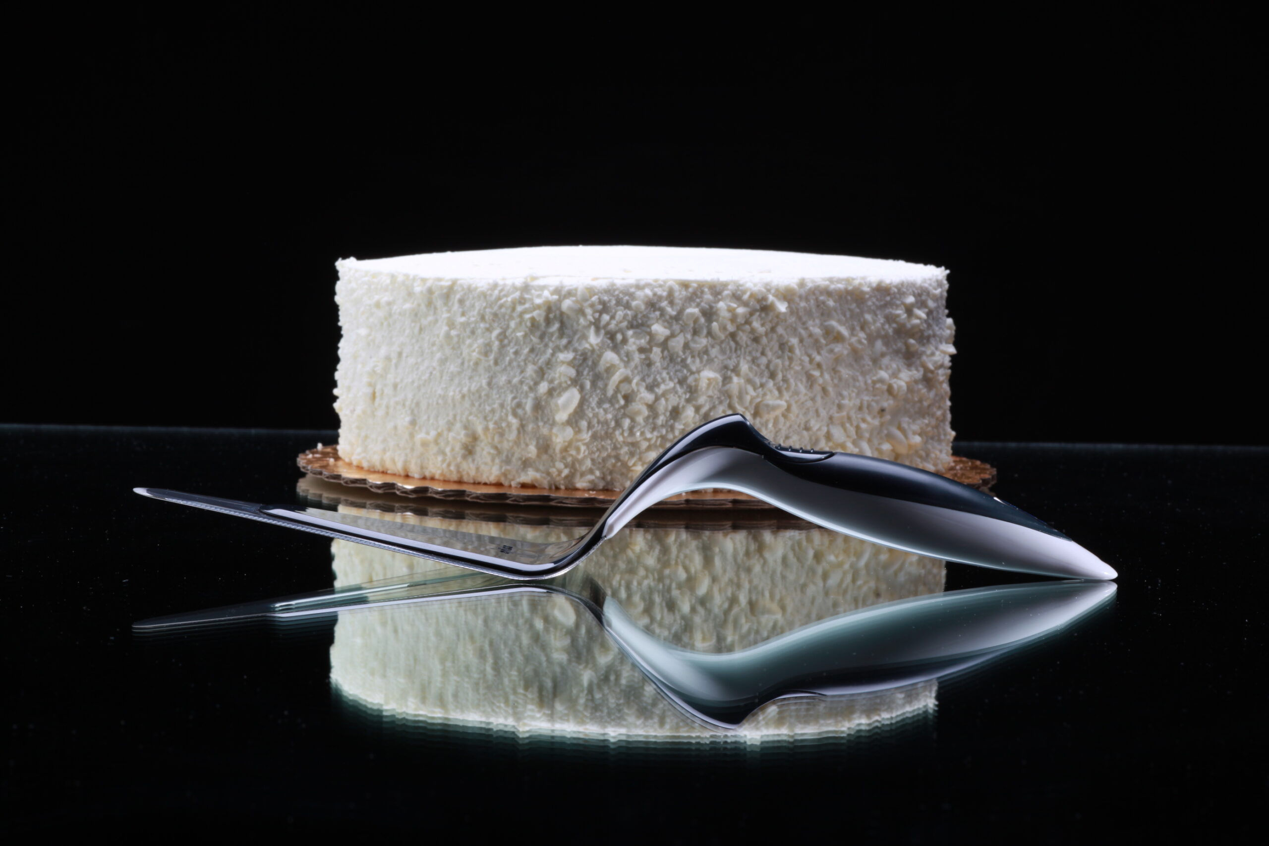 A white frosted round cake sits on a reflective black surface, with a shiny Zyliss fork and its reflection placed in front. The dark background highlights the cake and showcases the elegance of quality Swiss kitchen tools.